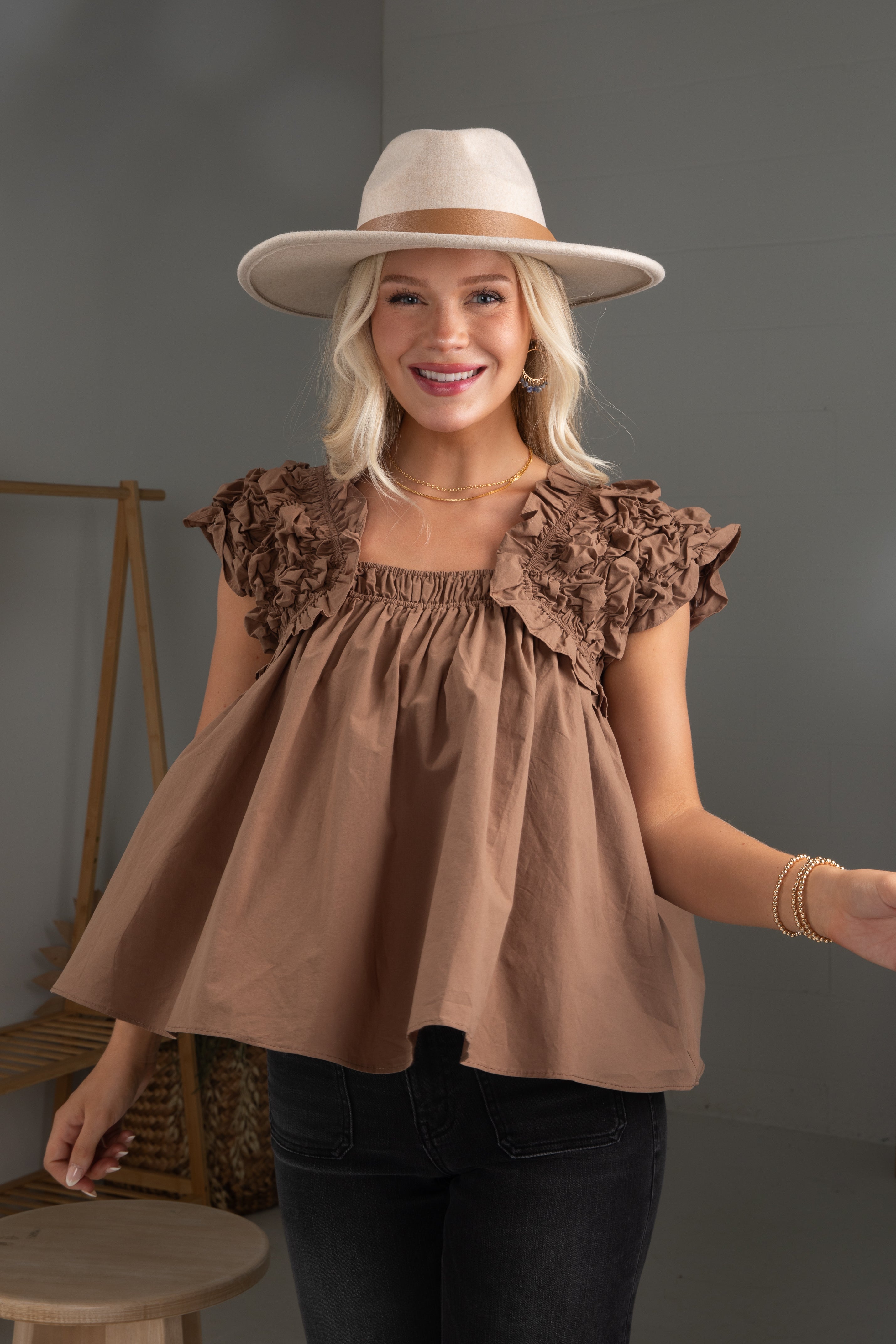 Woman wearing a brown top with ruffled shoulders and a white hat against a gray background