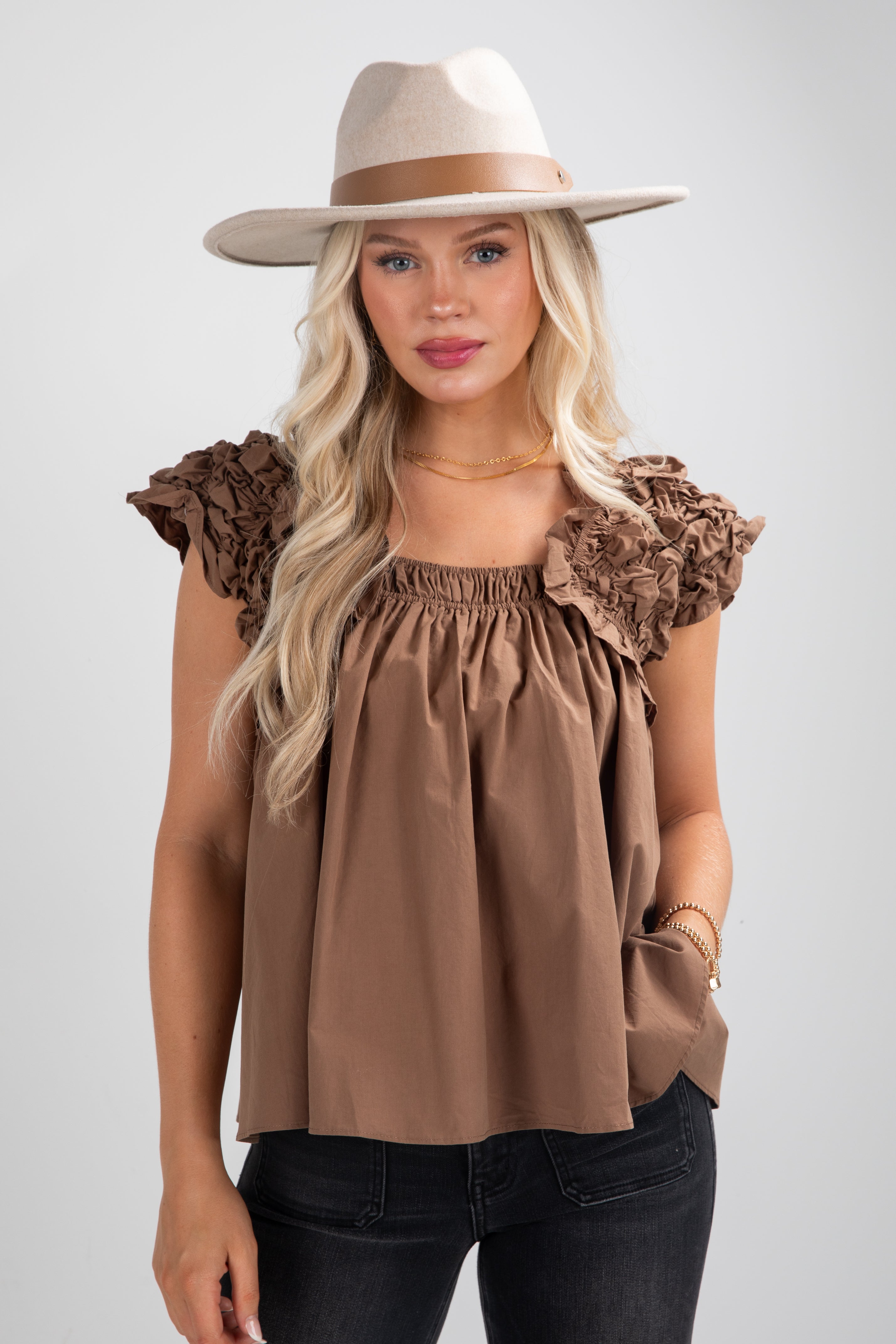 Woman wearing a brown top with ruffled sleeves and a beige hat on a white background