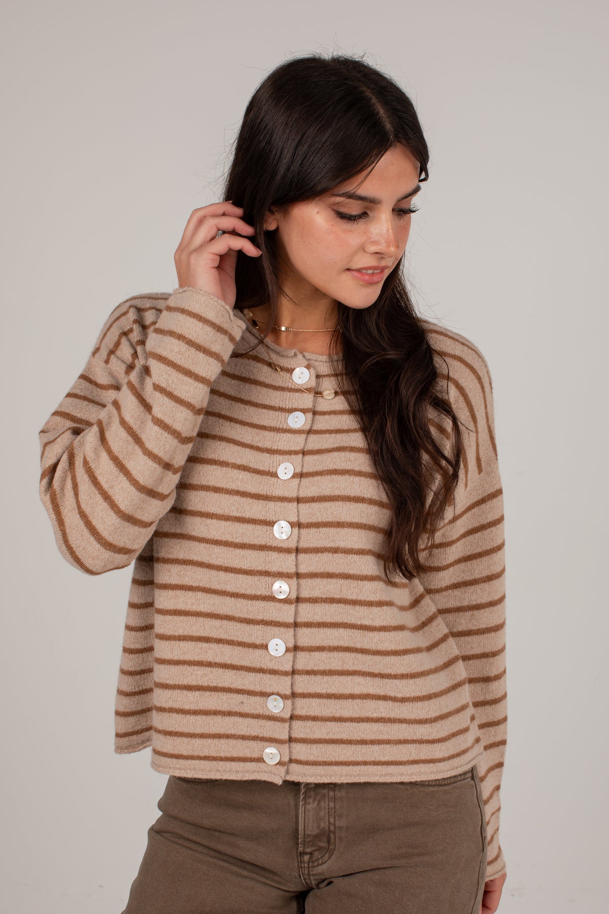 A woman wearing a cropped, long-sleeve knit cardigan with horizontal brown and beige stripes and white buttons down the front, paired with brown pants, against a plain light gray studio background.