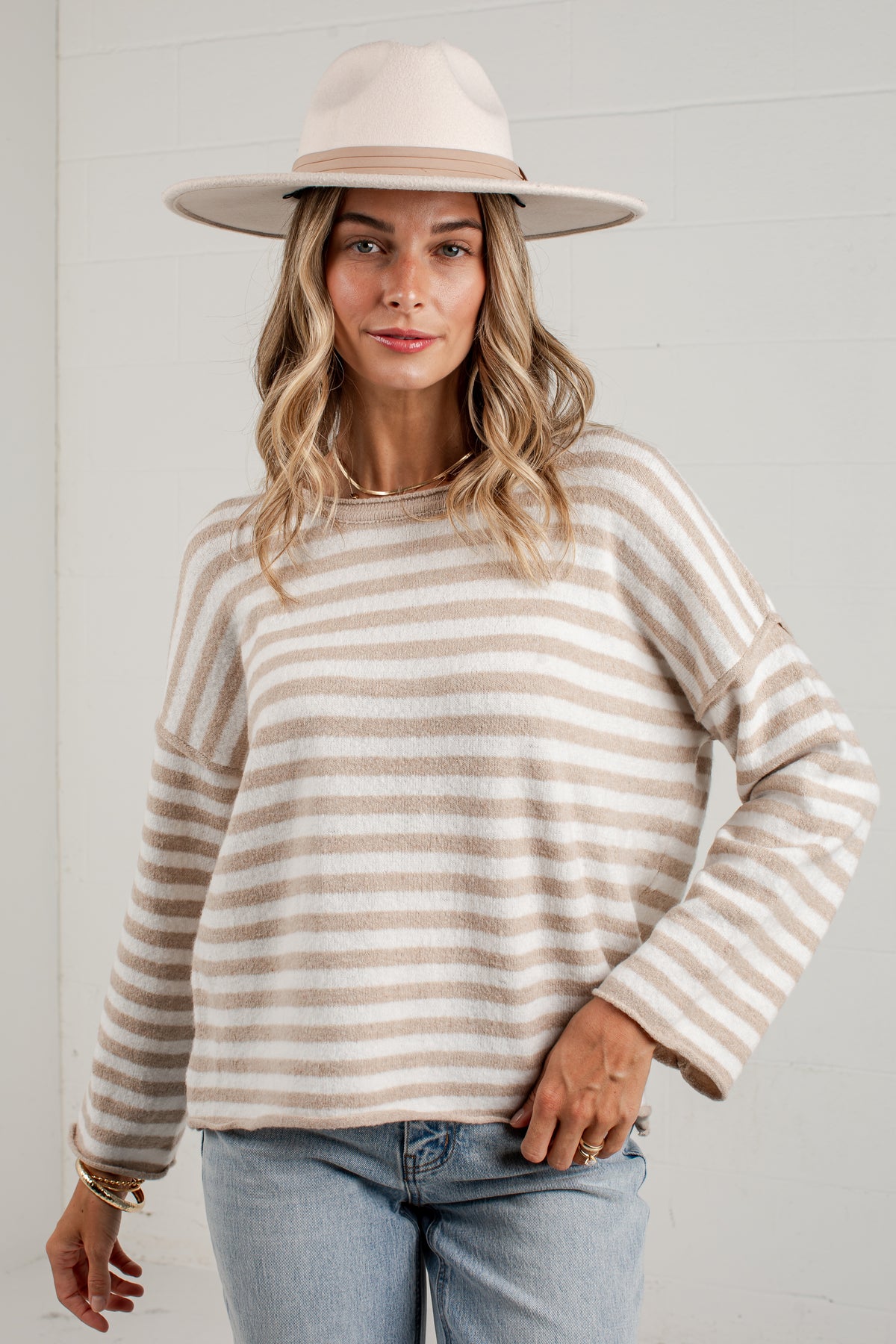 A woman wearing a beige and cream striped knit sweater, a wide-brimmed cream hat, and light blue jeans, standing against a white paneled wall.