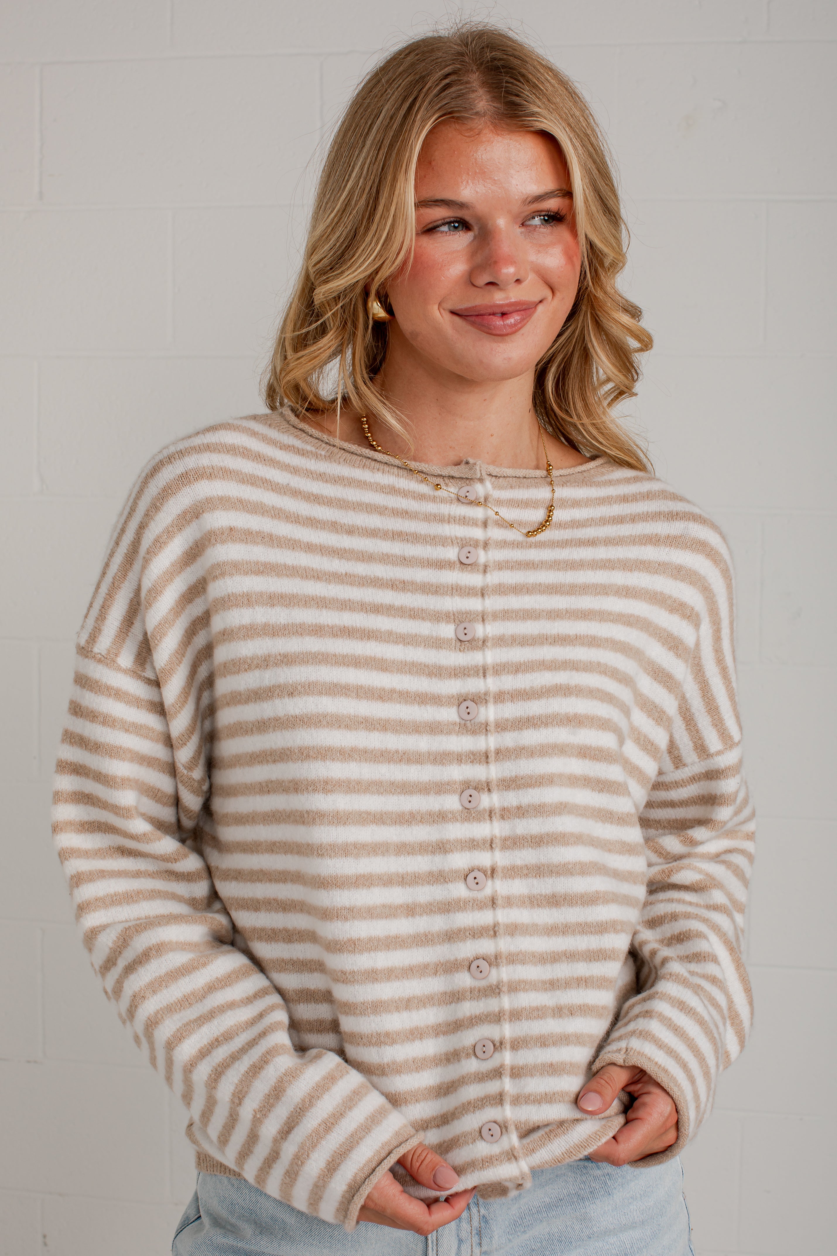 A woman wearing a beige and white horizontally striped button-up cardigan sweater, paired with light blue jeans, standing against a white brick wall.