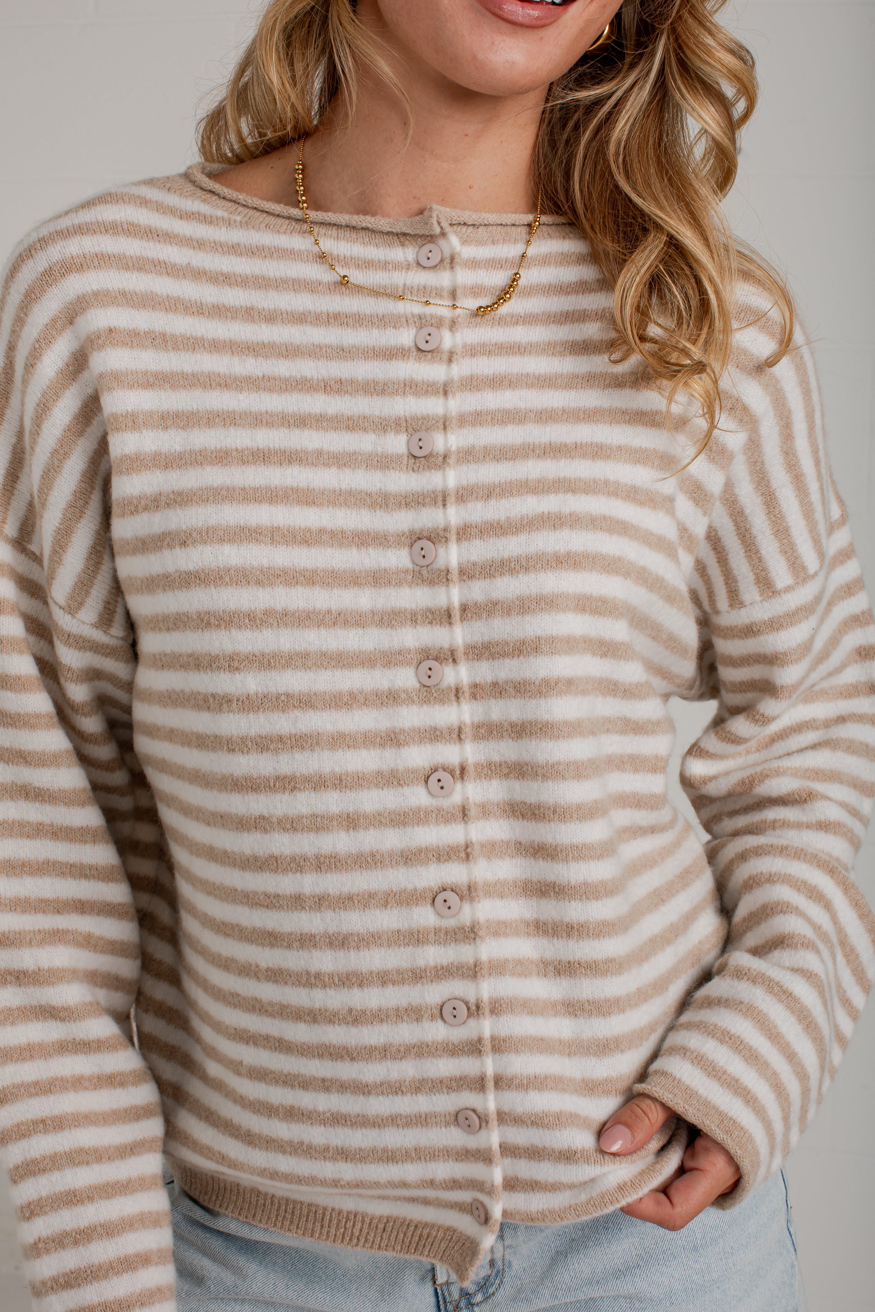 A woman wearing a beige and white horizontally striped button-up cardigan sweater, paired with light blue jeans and a gold necklace, against a neutral gray background.