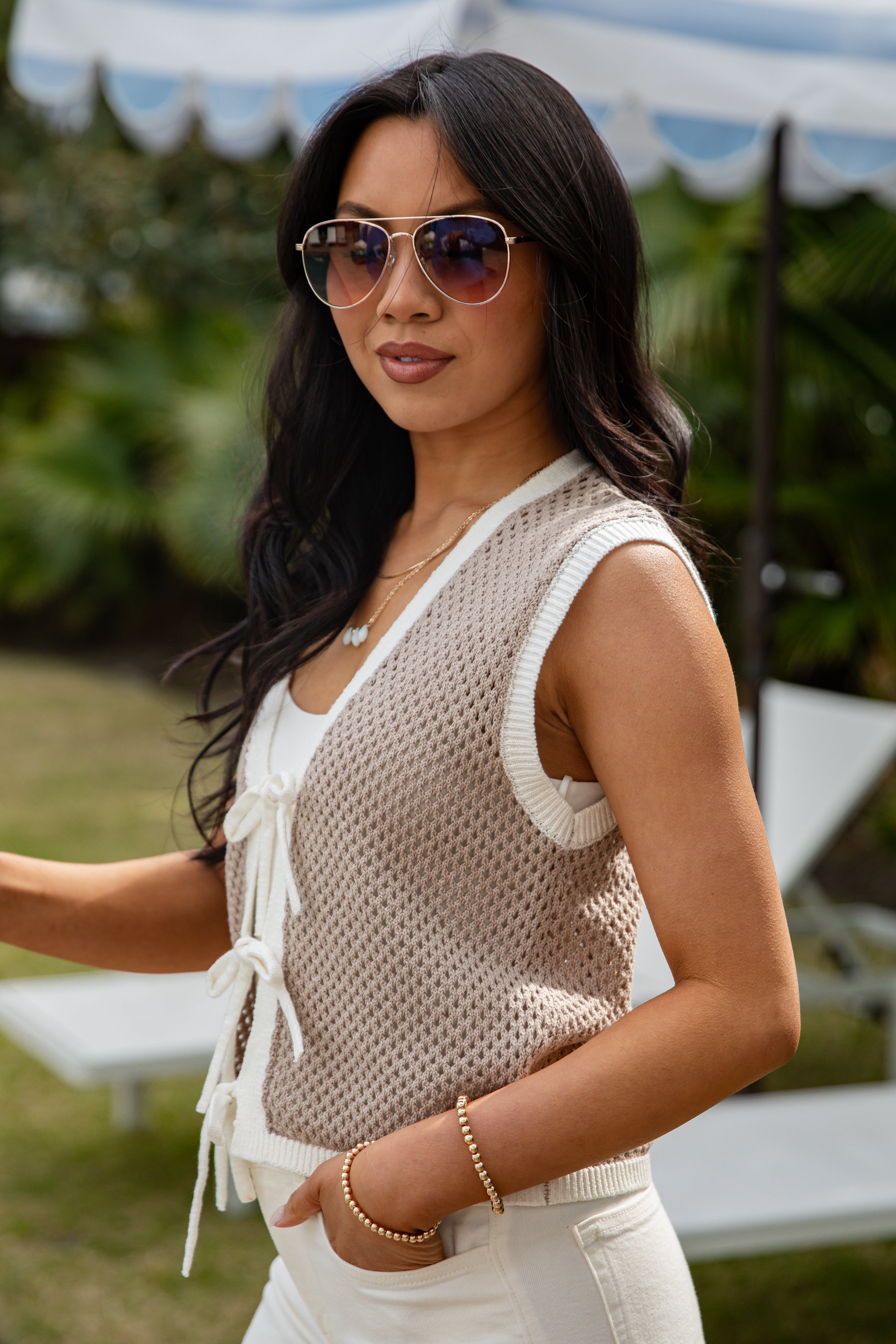 Woman wearing sunglasses and a beige sleeveless top with a blurred outdoor background