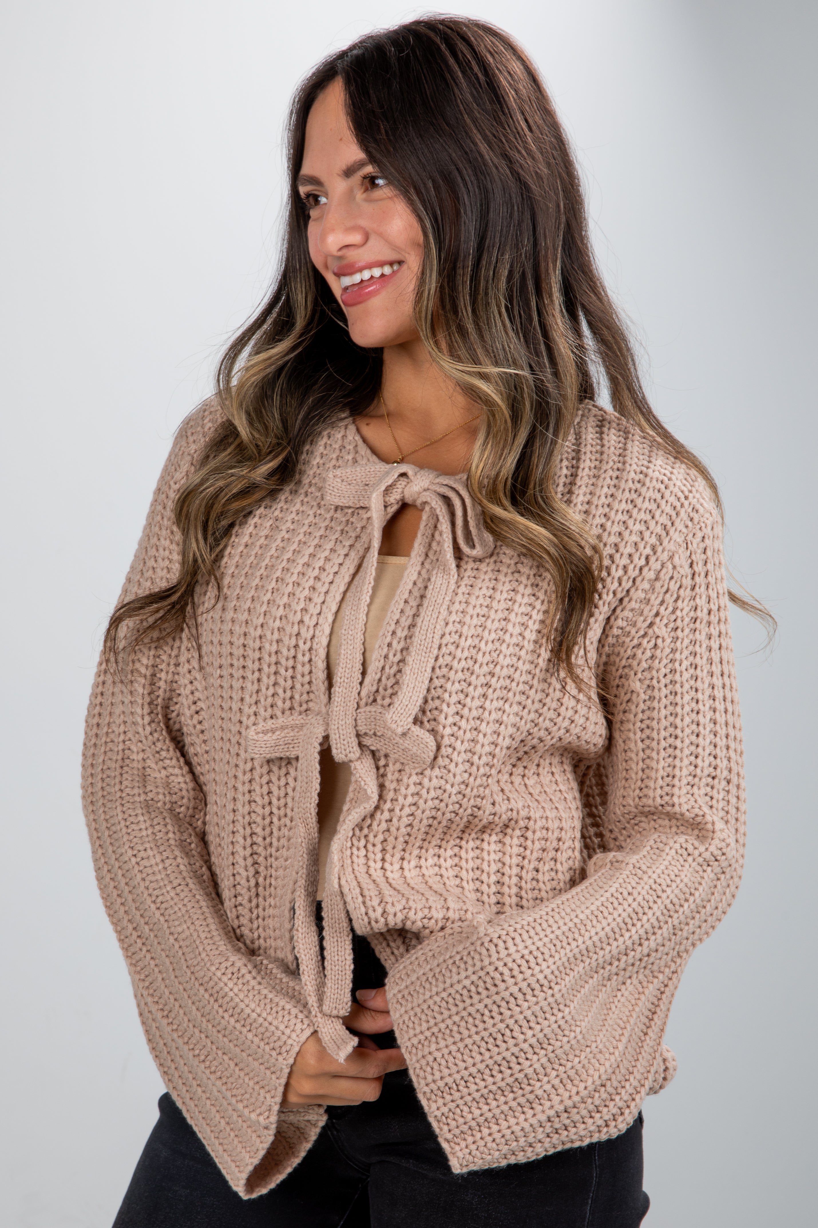 Woman wearing a beige knitted sweater with a tie detail on a plain background