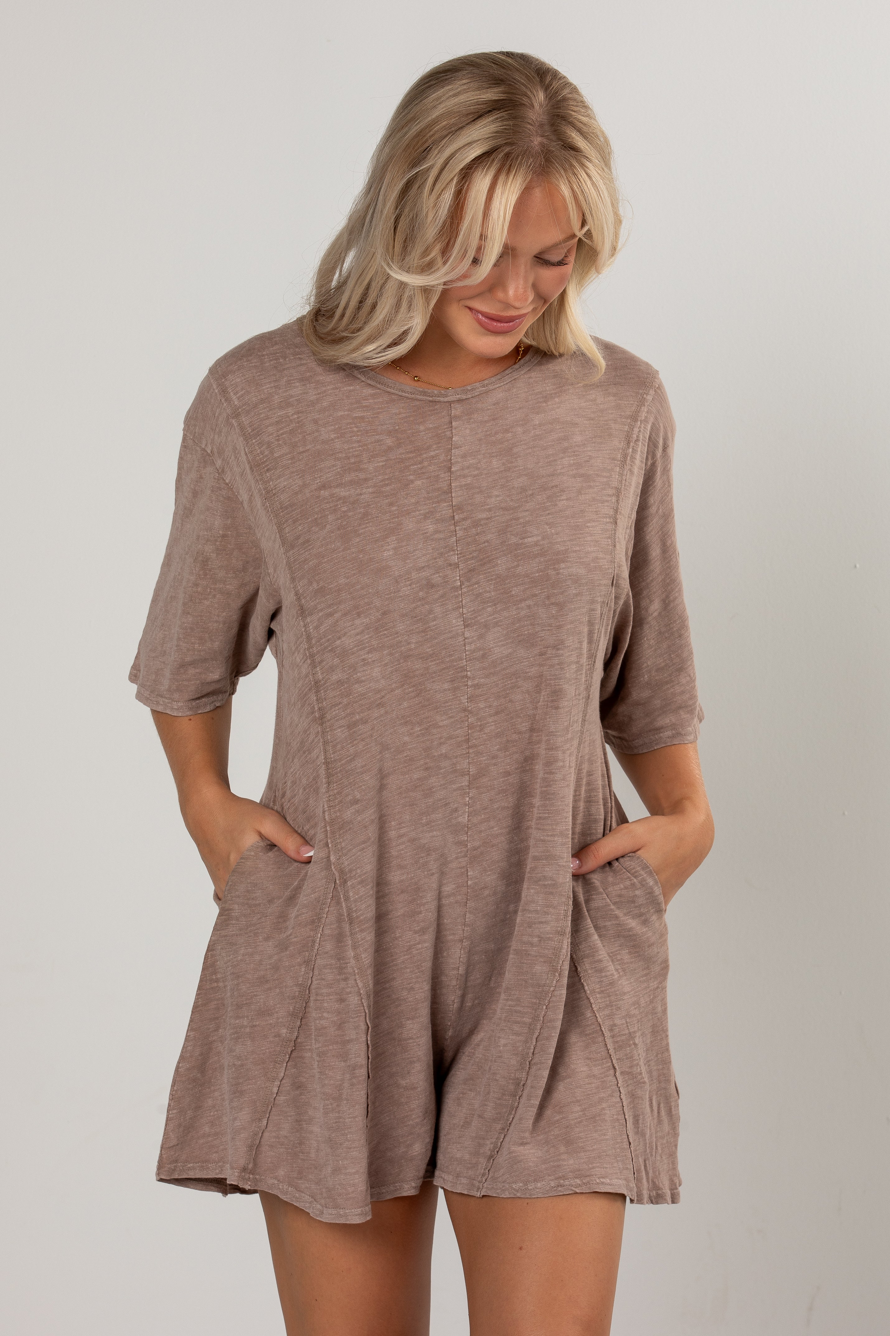 A woman wearing a short-sleeve, loose-fitting romper in a heathered taupe-brown color, standing against a light gray studio background.