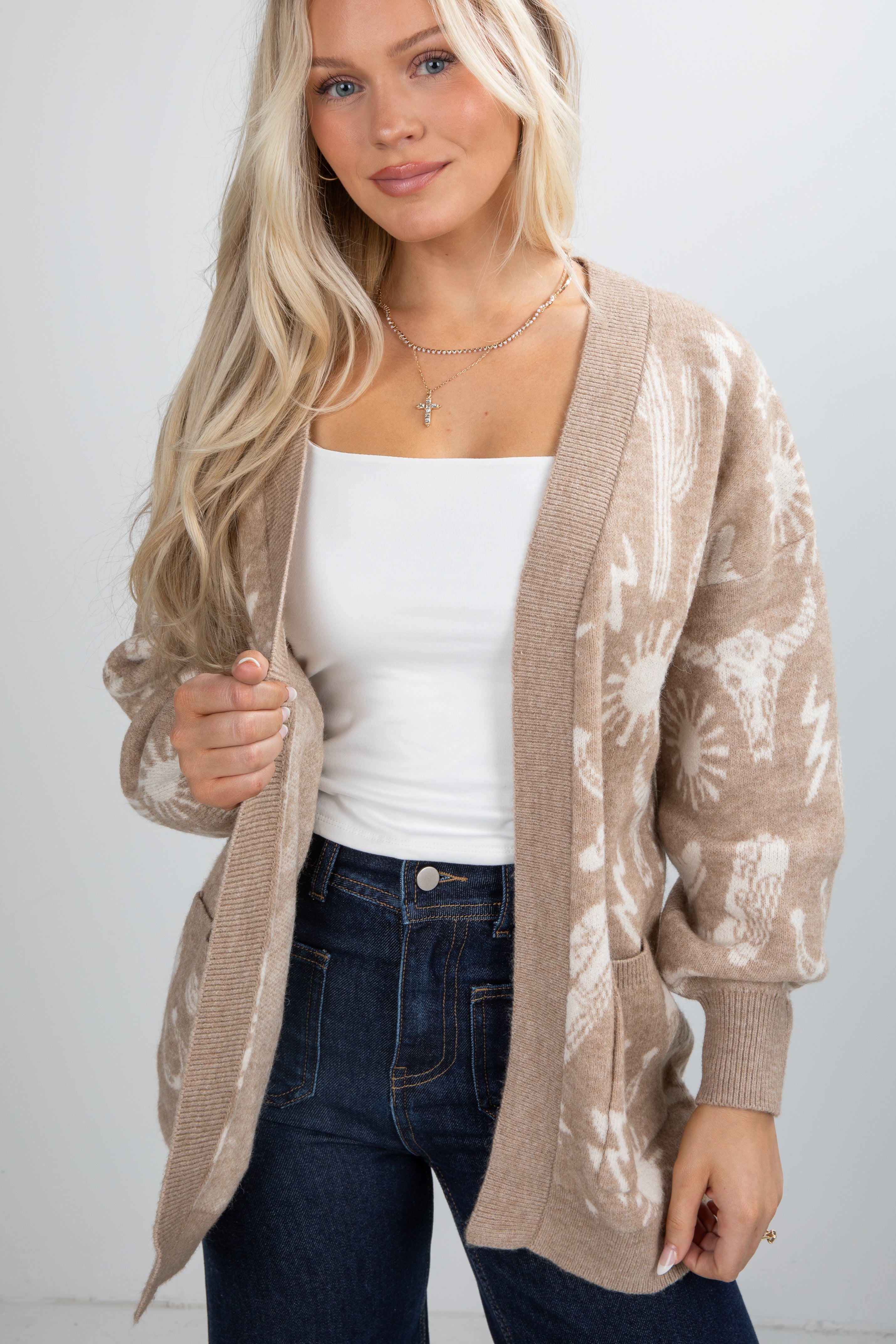 Woman wearing a beige patterned cardigan over a white top and dark jeans on a plain background