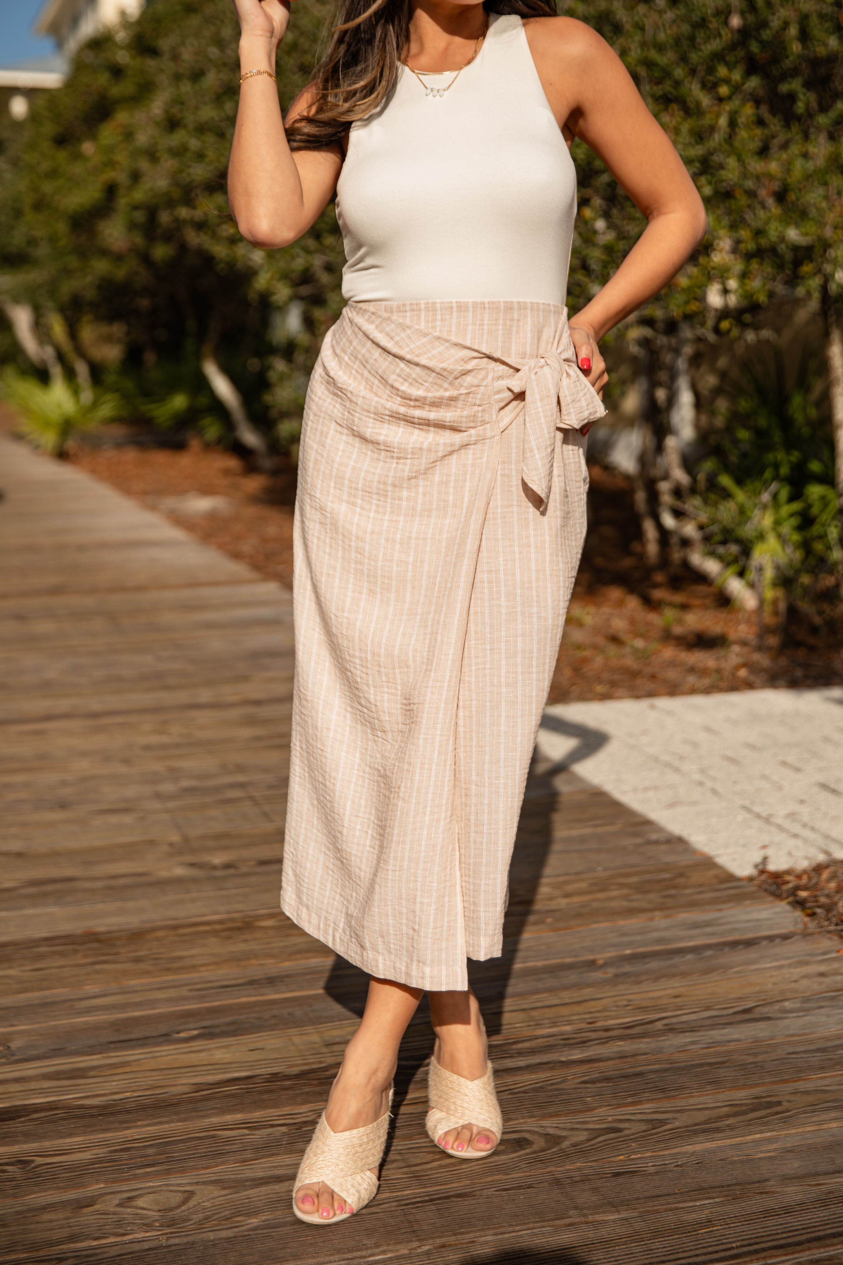 Woman wearing a white top and beige skirt on a wooden path with greenery in the background