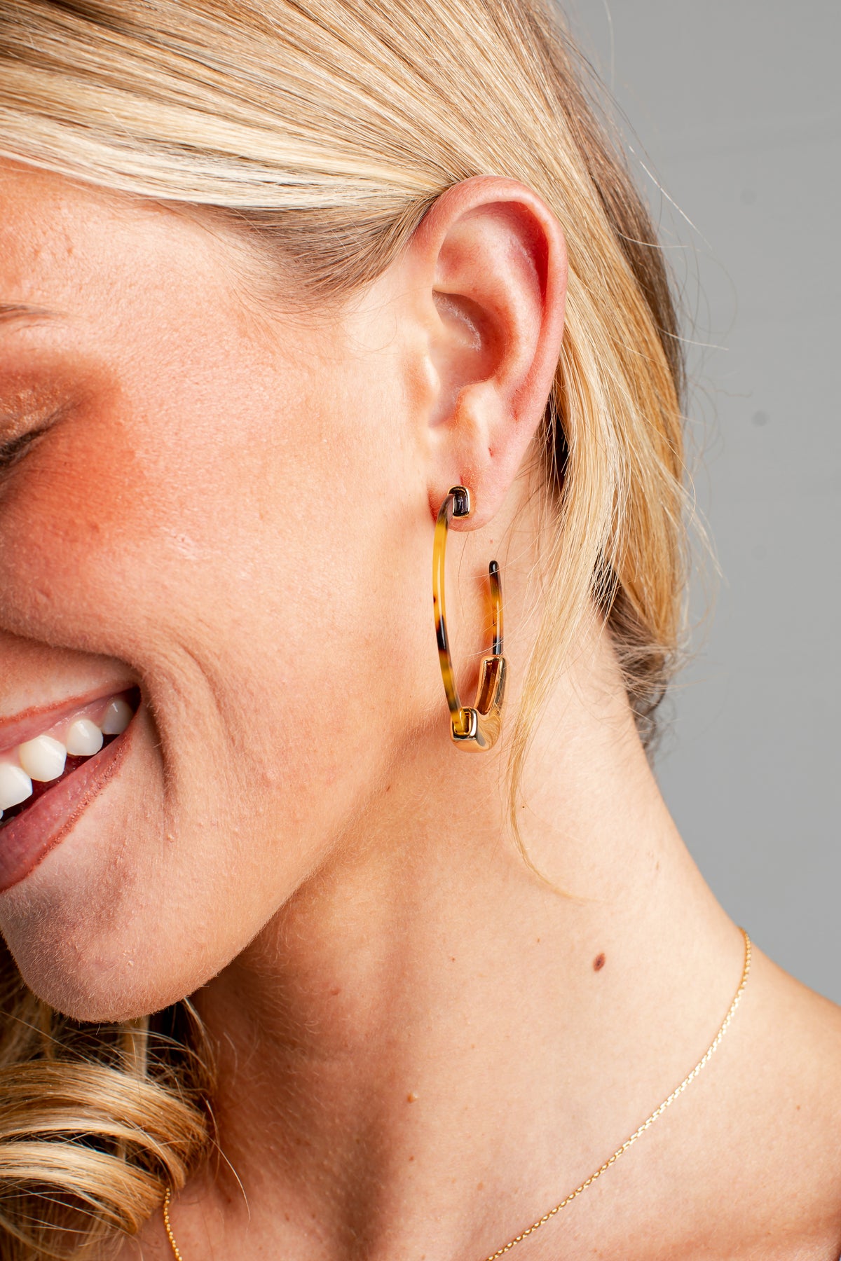 A close-up of a smiling blonde woman wearing a pair of gold hoop earrings with tortoiseshell-style brown and black mottled detailing.