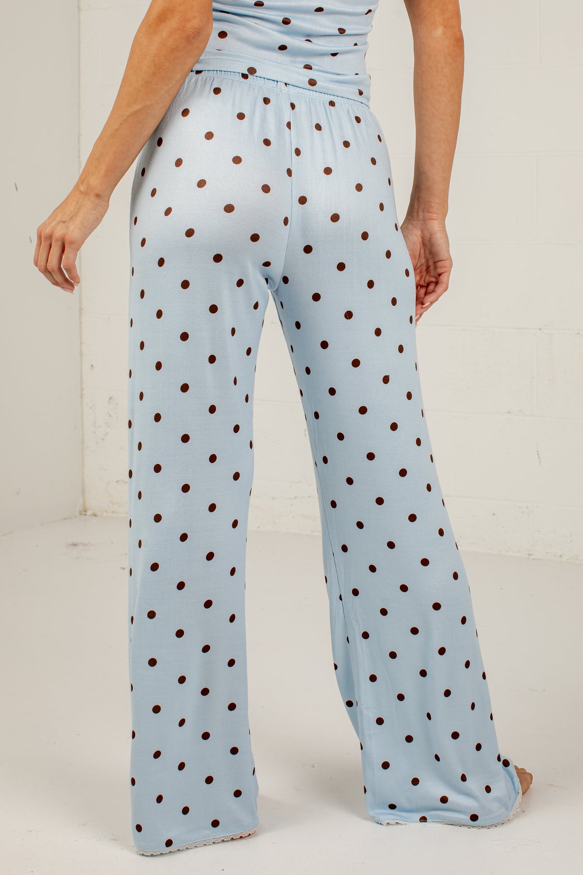 A person shown from the back wearing light blue wide-leg pants with an allover dark brown polka dot pattern, standing against a white wall.