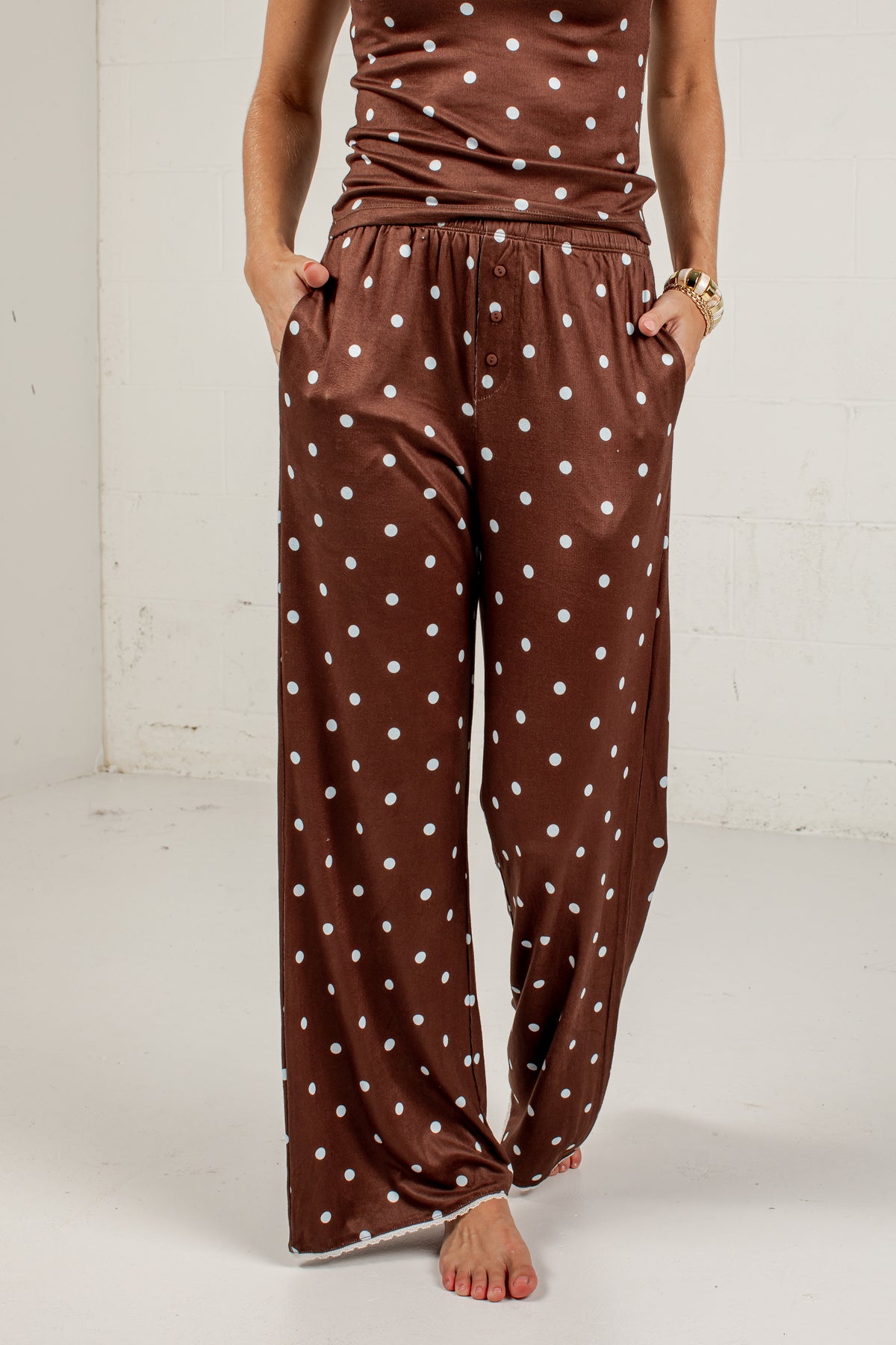 A person wearing brown wide-leg pants with a white polka dot pattern, standing barefoot against a white studio wall.