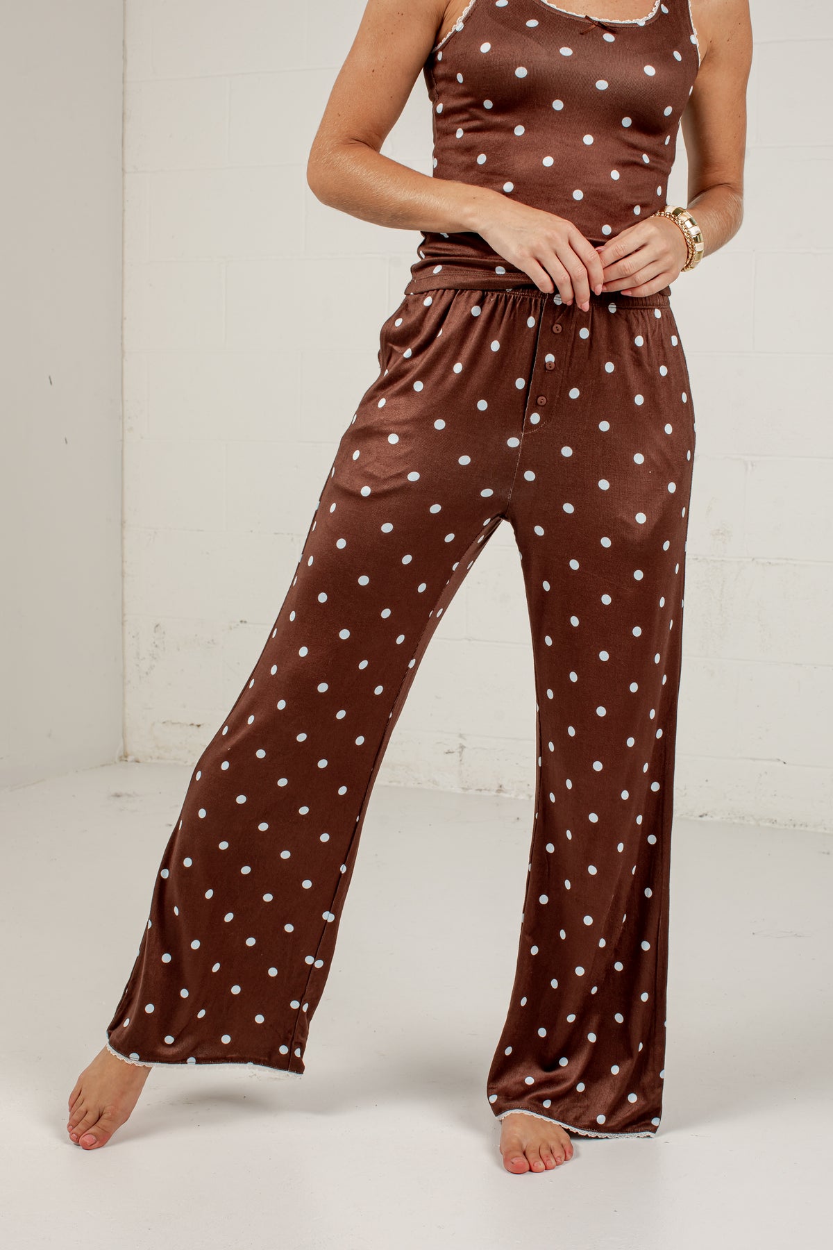 A woman modeling a brown stretchy two-piece pajama set with white polka dots, featuring a sleeveless top and wide-leg pants, against a white studio background.