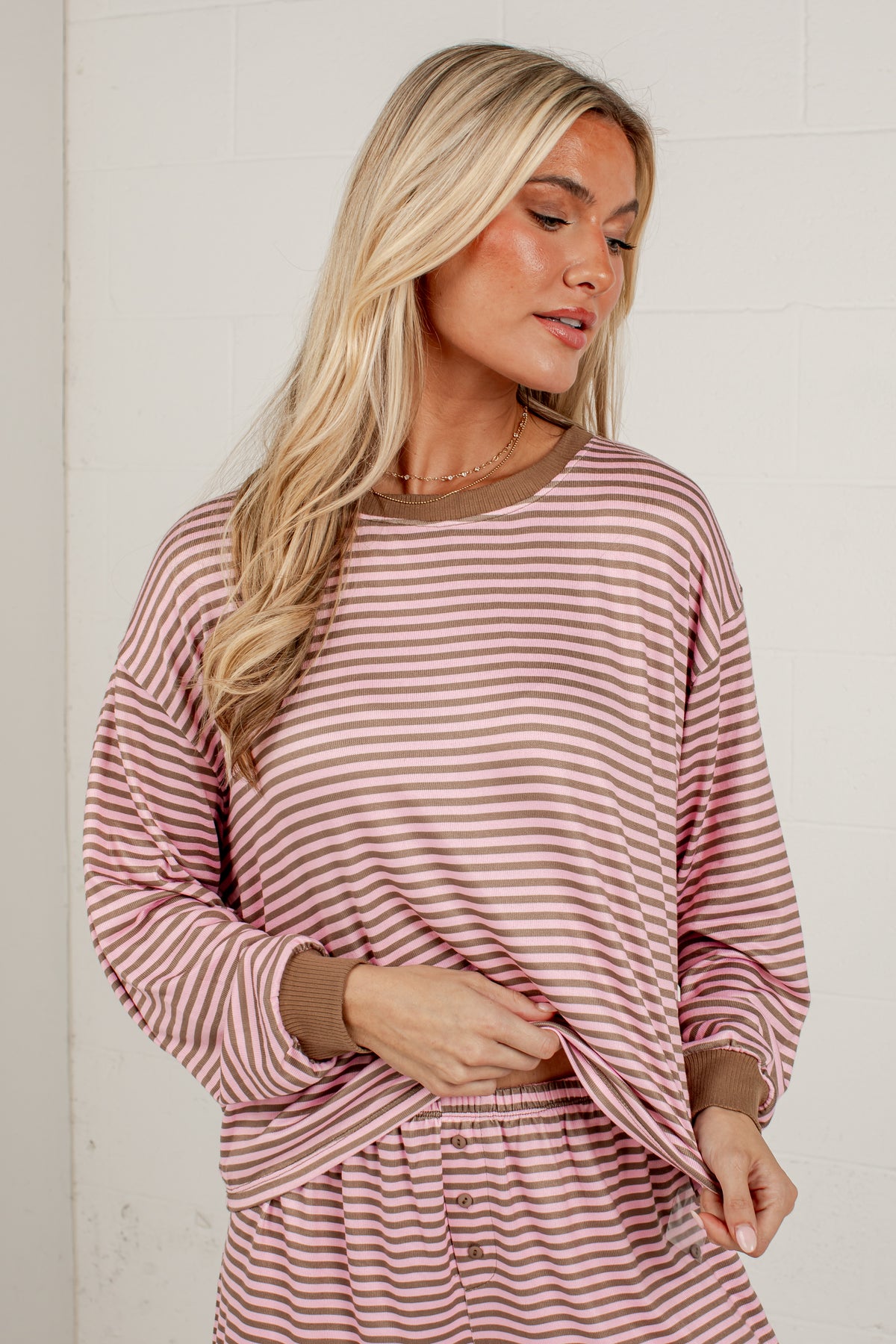 A woman wearing a dusty rose and brown striped long-sleeve pajama top with matching bottoms, standing against a white brick wall.