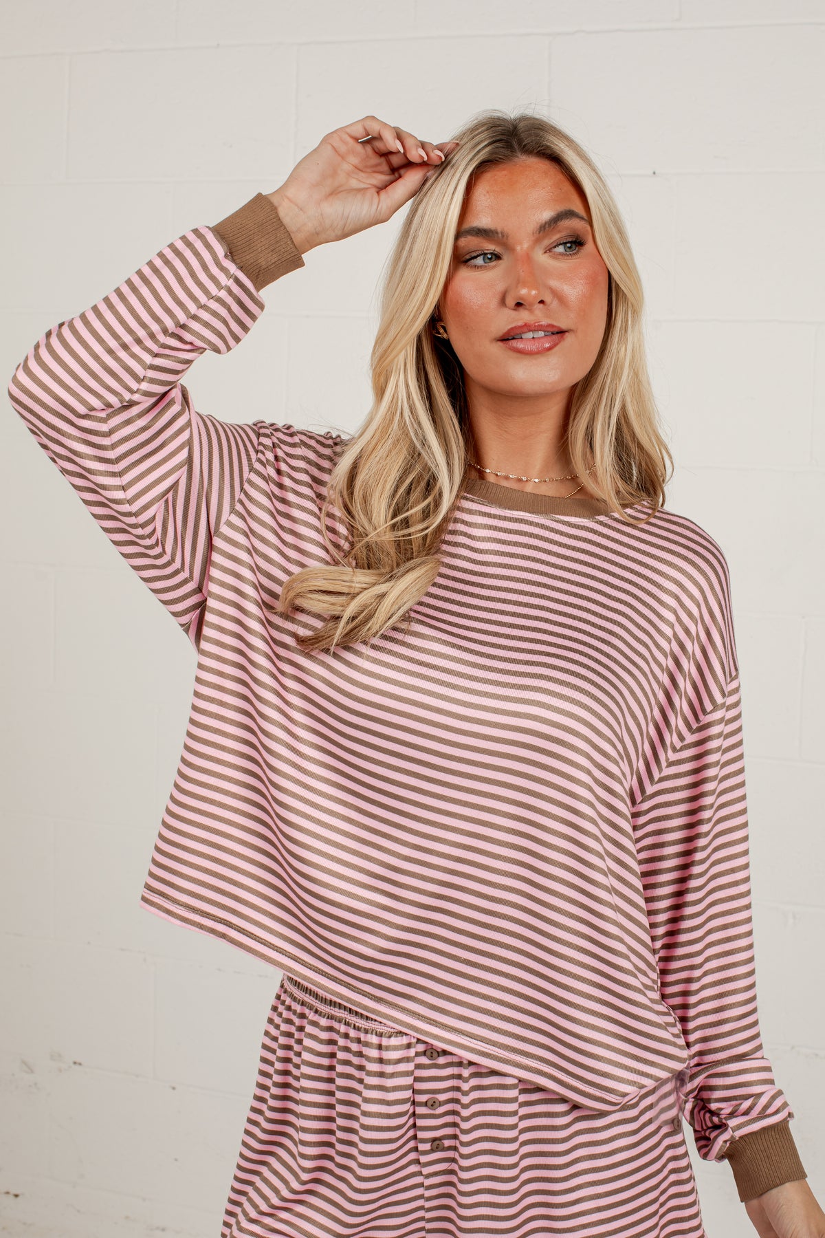 A woman modeling a long-sleeve striped top with a dusty rose and white pattern, featuring solid brown ribbed cuffs and collar, set against a white brick wall background.