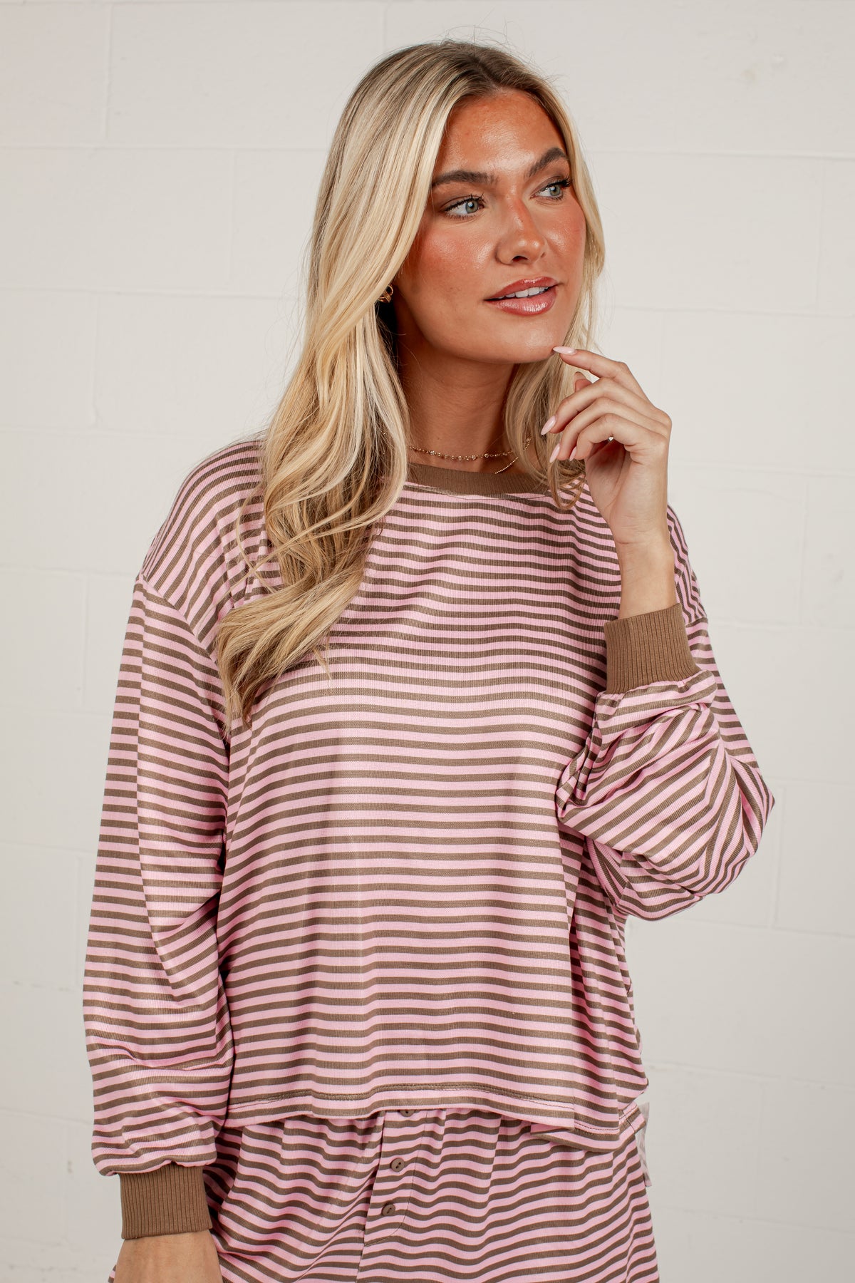 A woman wearing a long-sleeve striped lounge top in dusty pink and brown, with matching striped bottoms partially visible. She is posing against a light off-white brick wall.