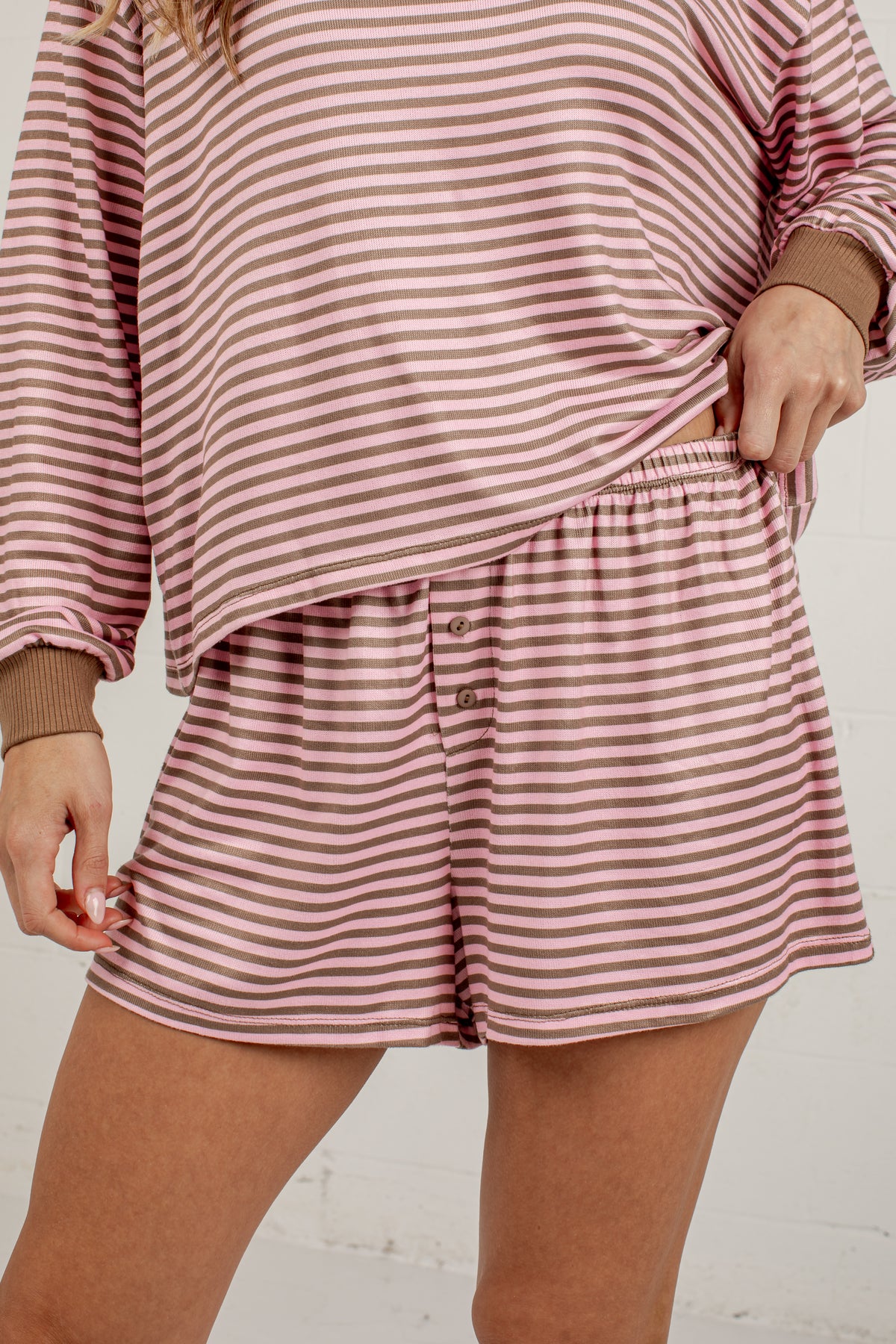 A close-up of a person wearing a matching two-piece loungewear set with horizontal pink and white stripes, featuring a long-sleeve top and shorts with brown ribbed cuffs and waistband.