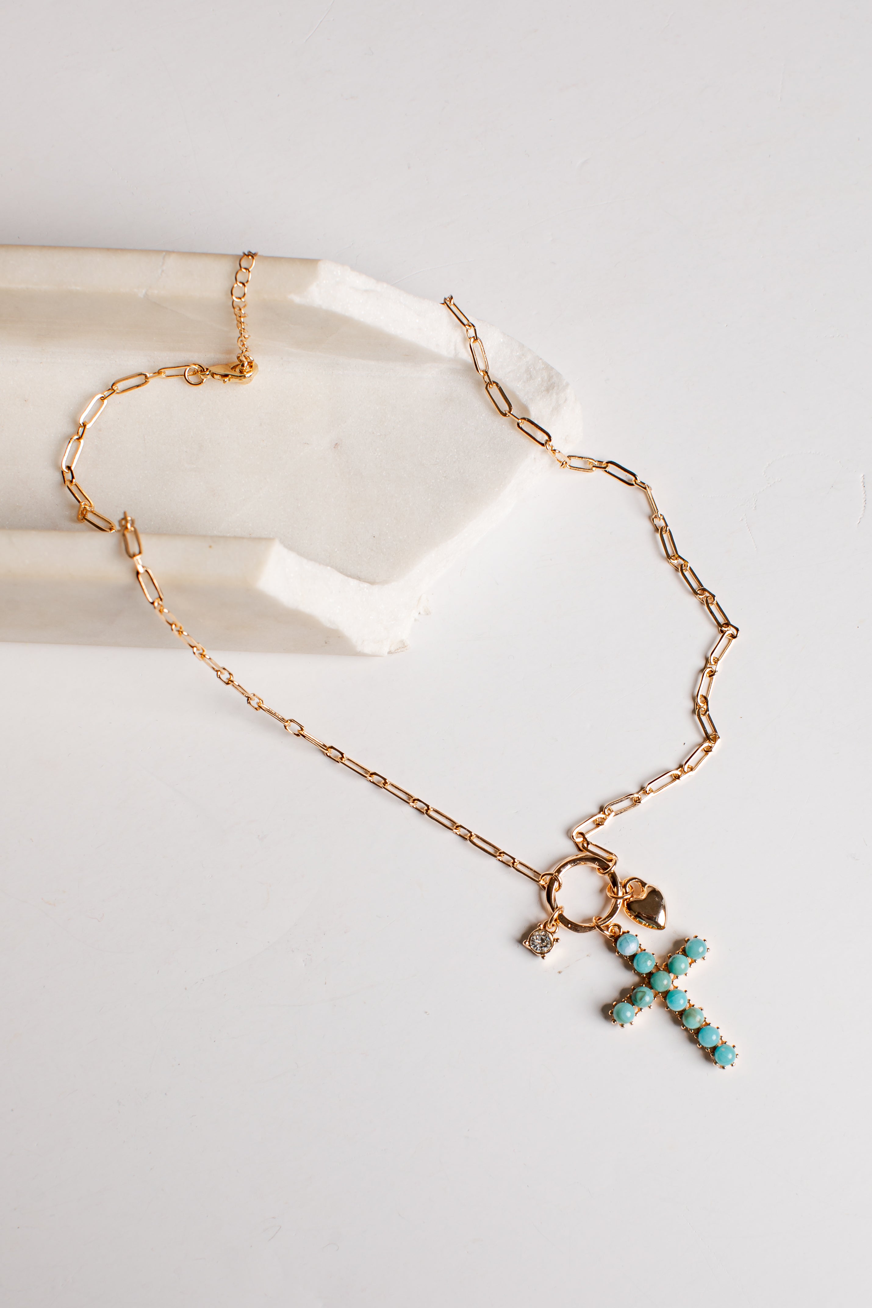 A gold chain necklace with a cross-shaped pendant studded with turquoise blue beads, displayed on a white surface next to a white stone block.