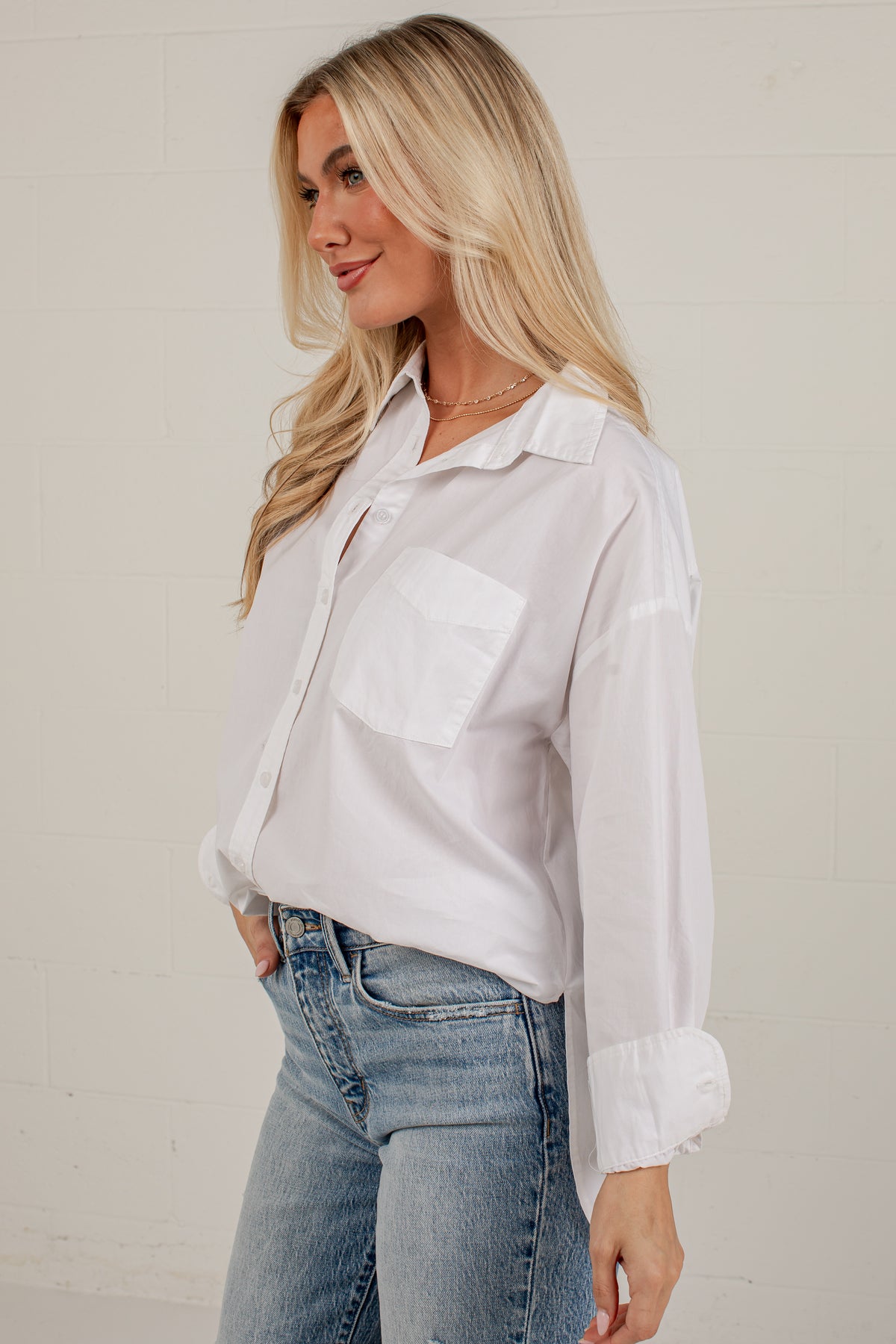A woman wearing a white long-sleeve button-up shirt with a chest pocket and light blue jeans, standing against a light off-white brick wall.