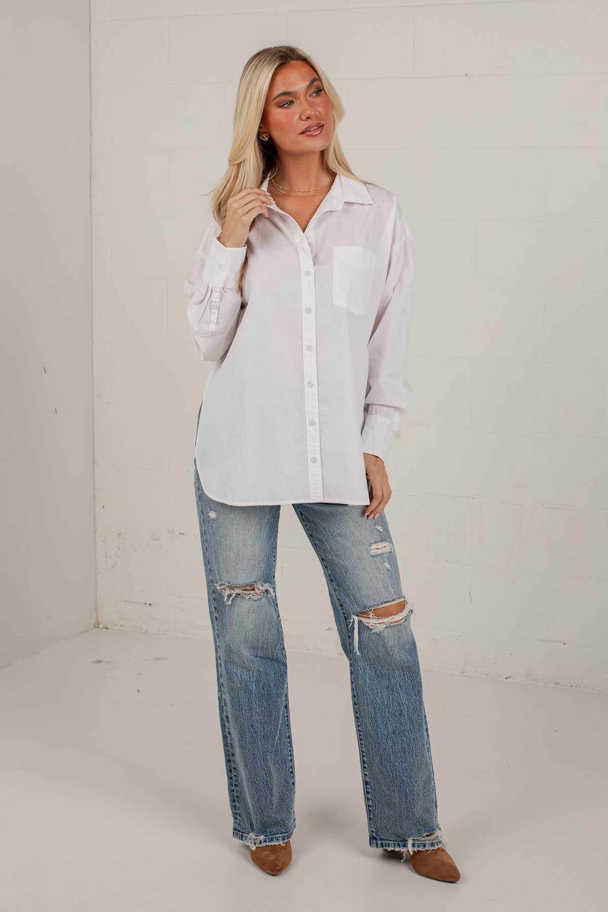 A woman modeling a white long-sleeve button-up shirt with a chest pocket, paired with distressed blue jeans and brown pointed-toe flats, standing against a white studio wall.