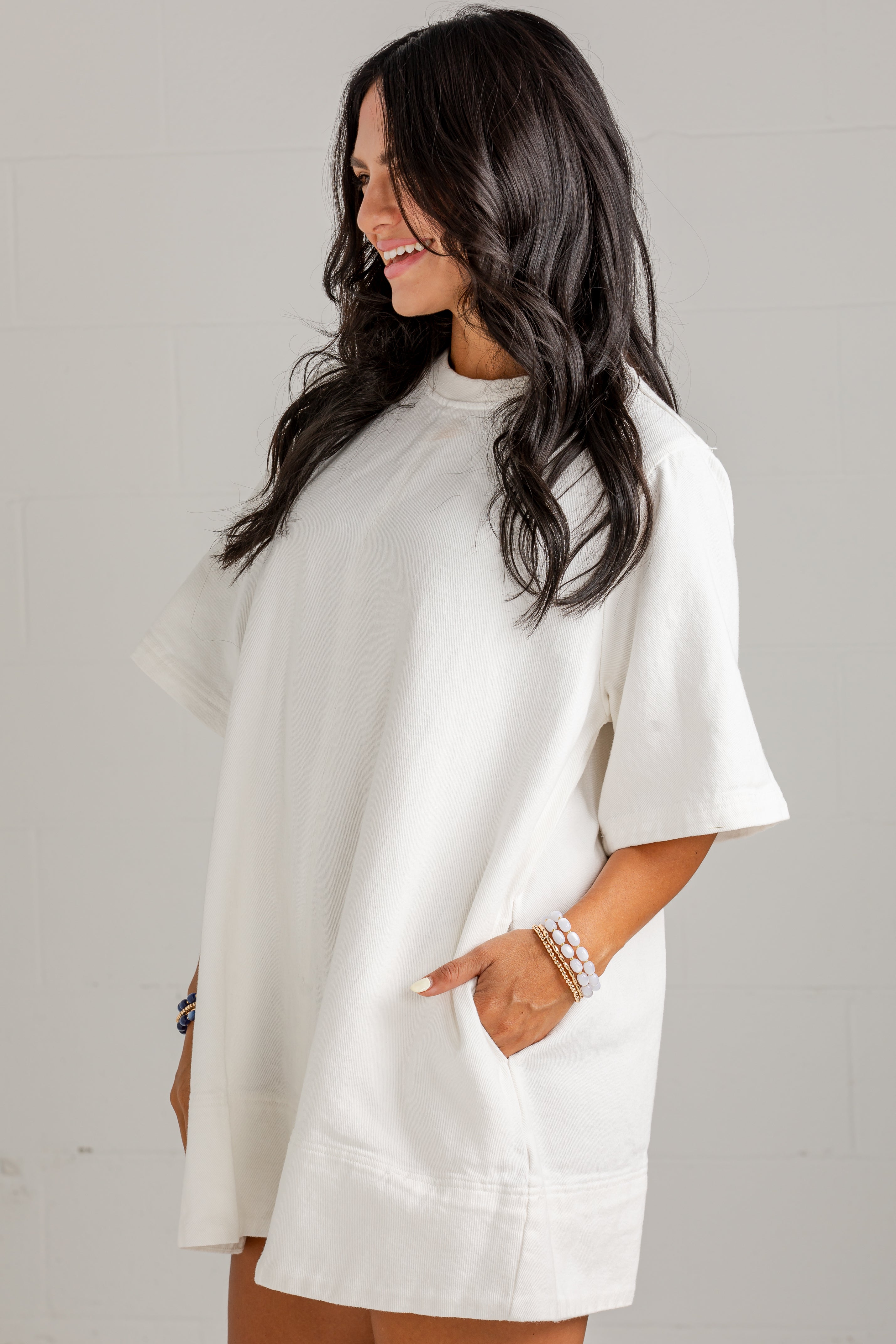 A woman wearing an oversized white short-sleeve tunic top with side pockets, standing against a light gray wall.