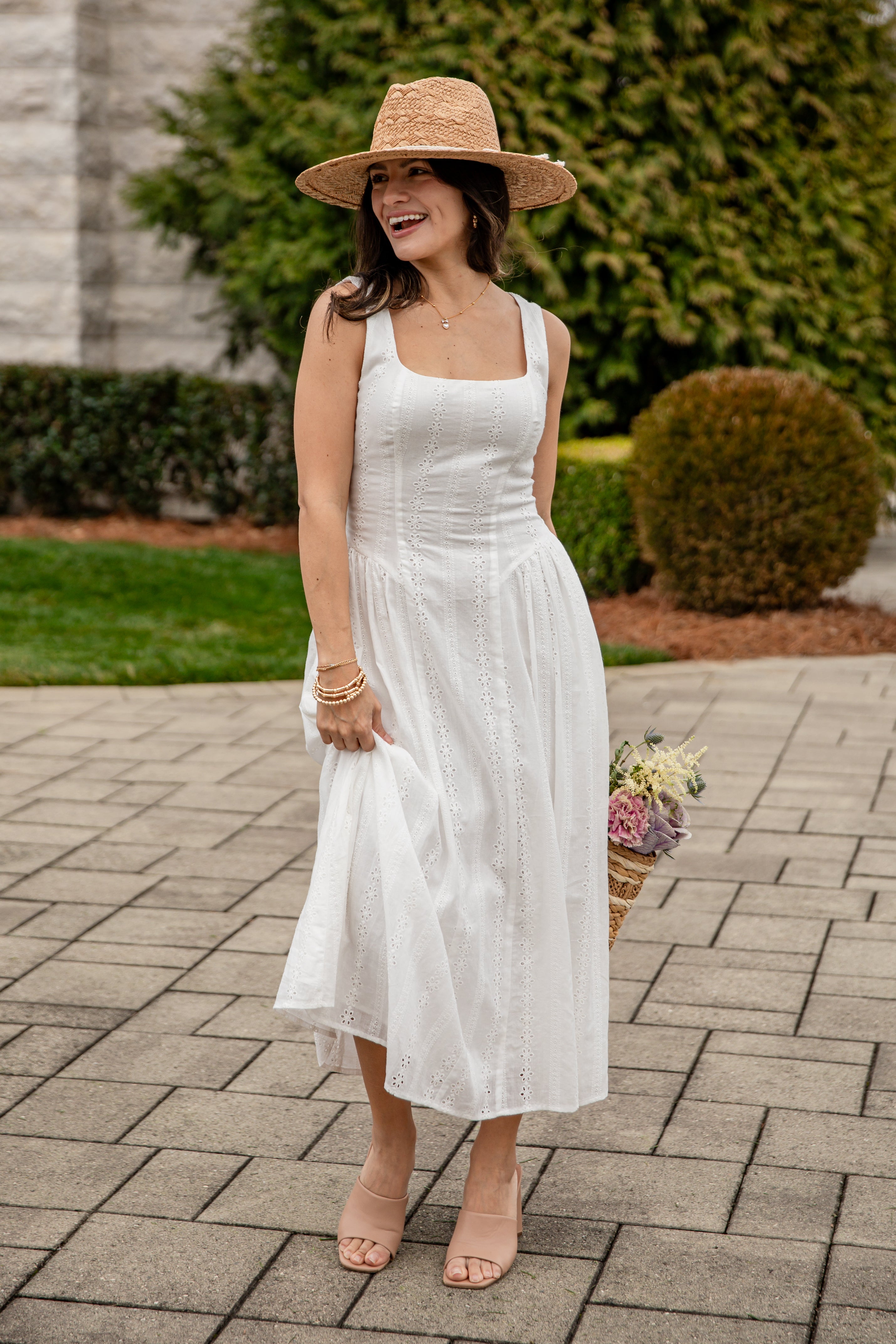 Woman in a white dress and straw hat standing on a paved walkway with greenery in the background