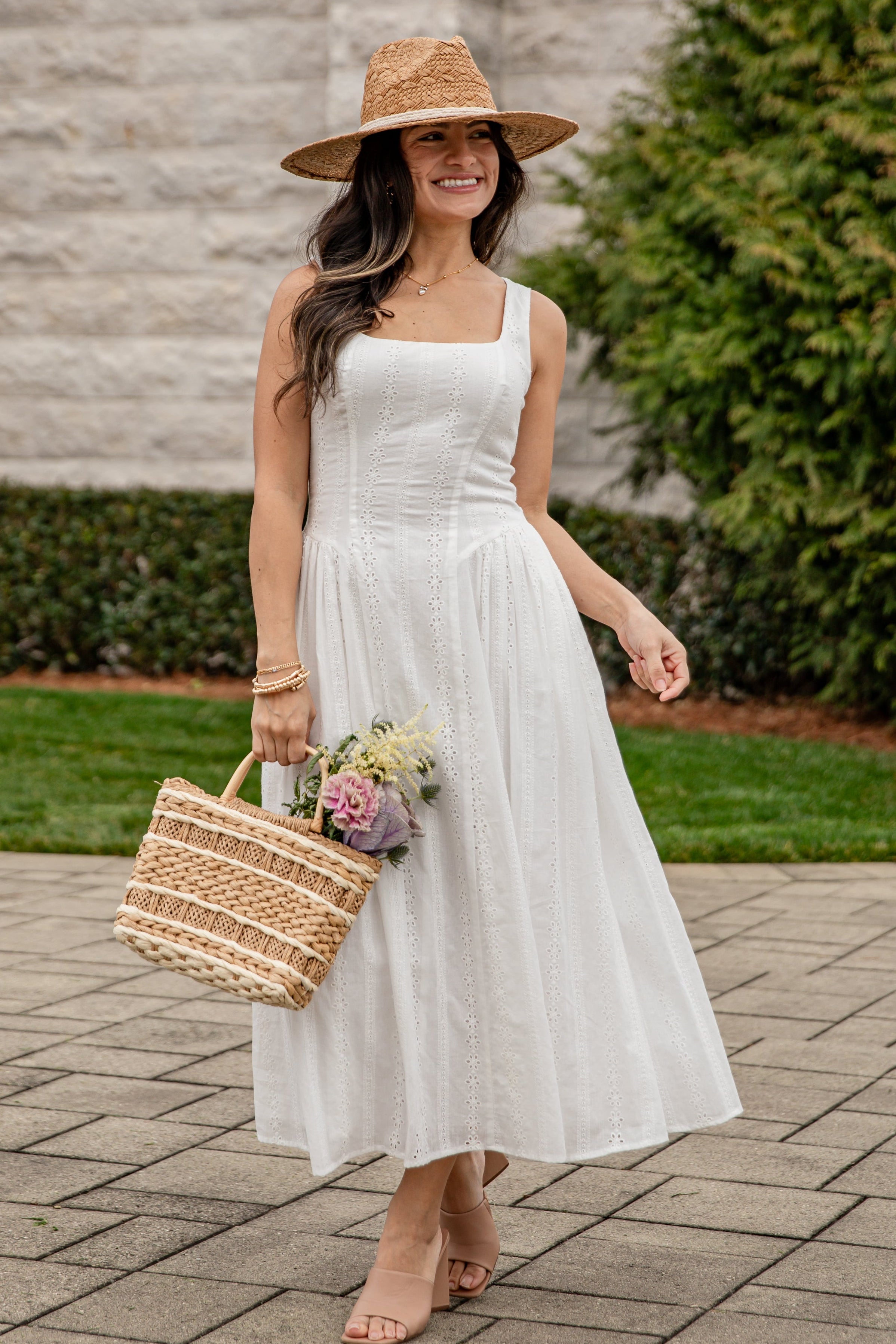 Woman in a white dress and straw hat holding a basket and flowers outdoors.