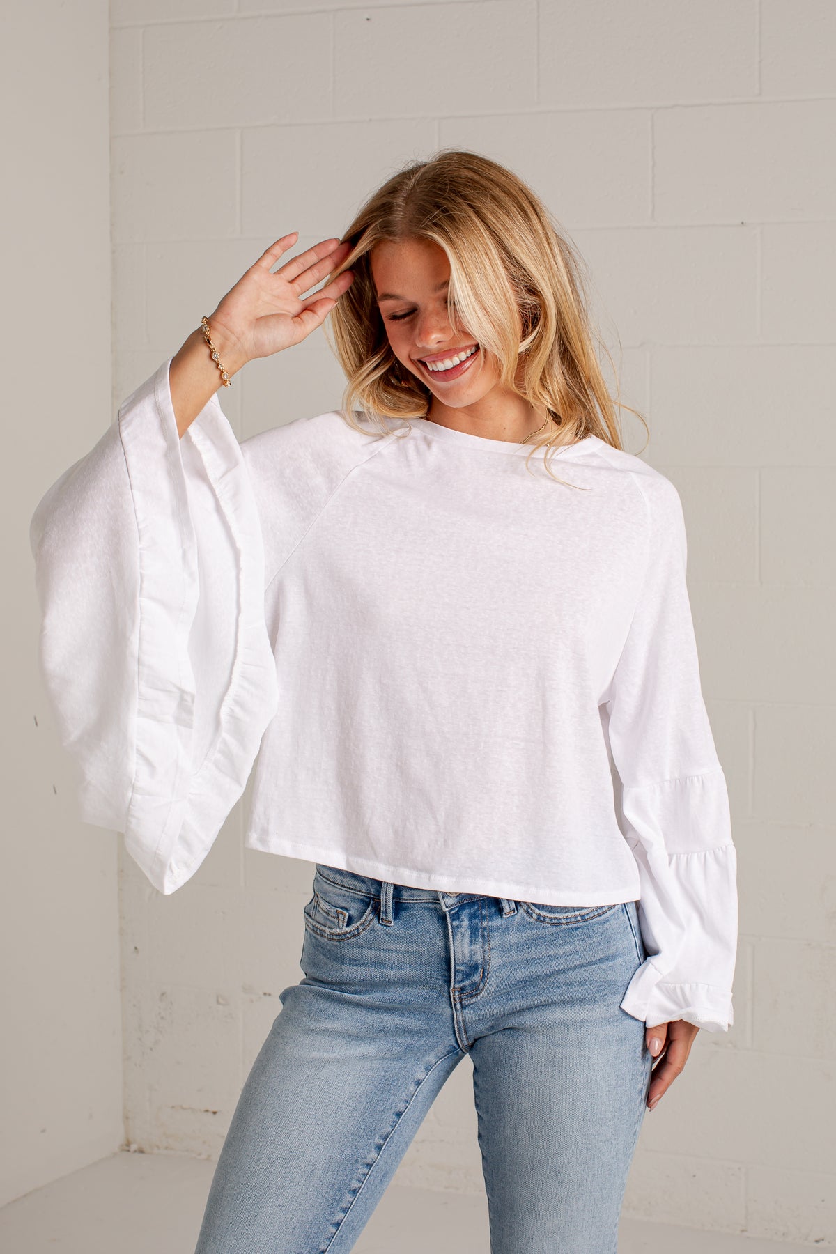 A woman wearing a white long-sleeve crop top with dramatic ruffled bell sleeves and light-wash blue jeans, standing against a white brick wall.