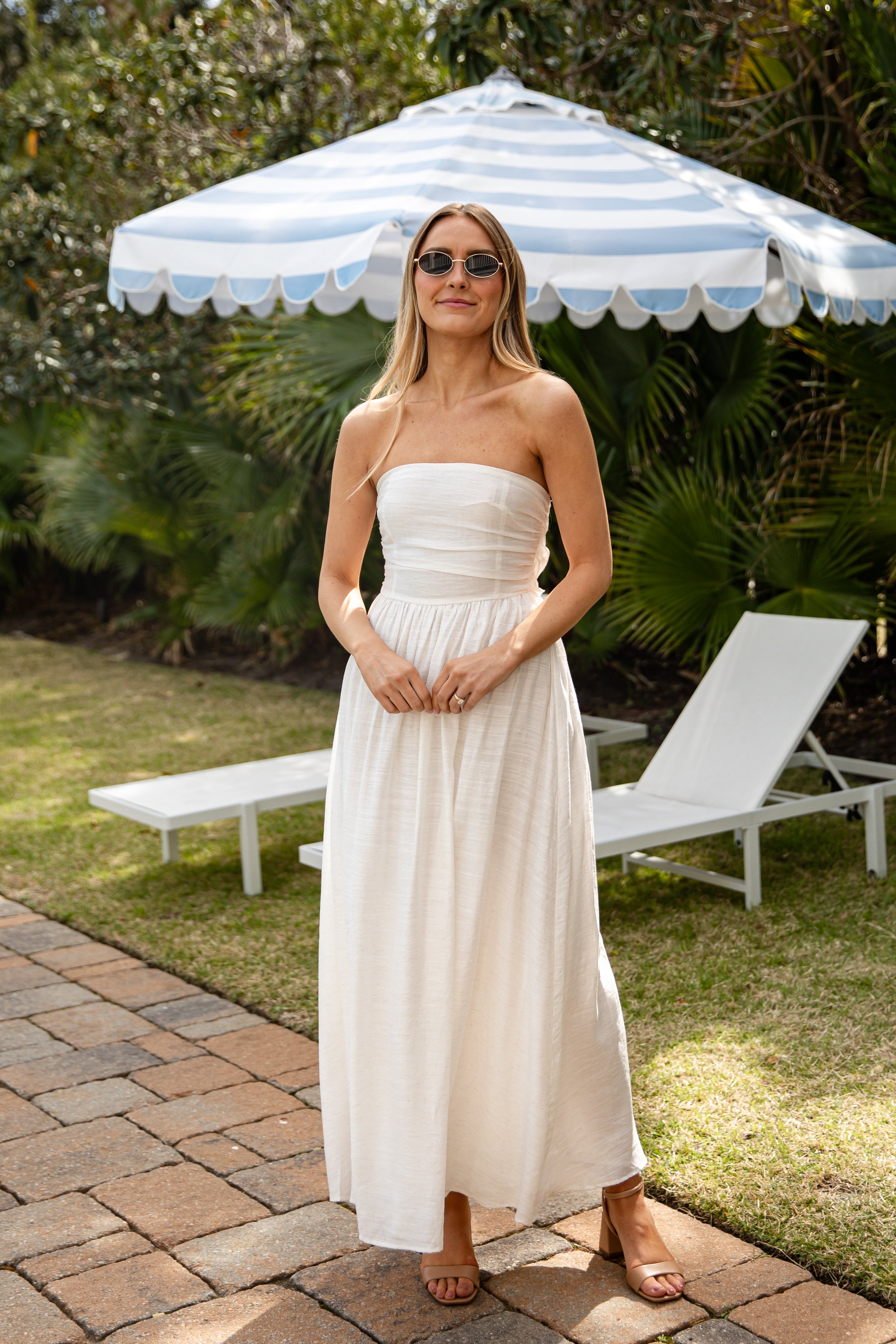 Woman in a strapless white dress standing under a striped umbrella in a garden setting.