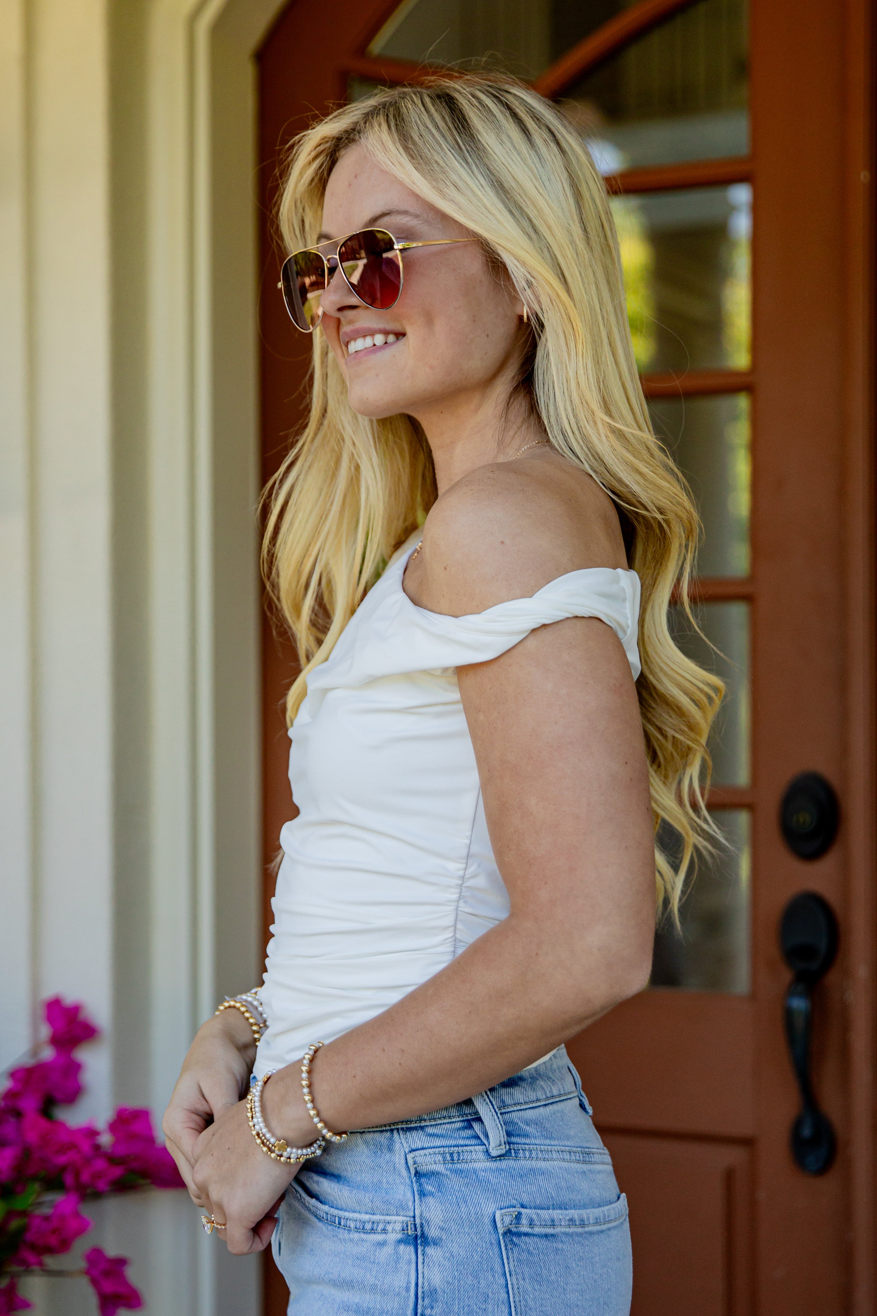 A blonde woman wearing a white off-the-shoulder top, light blue jeans, and round sunglasses stands in front of a brown wooden door.