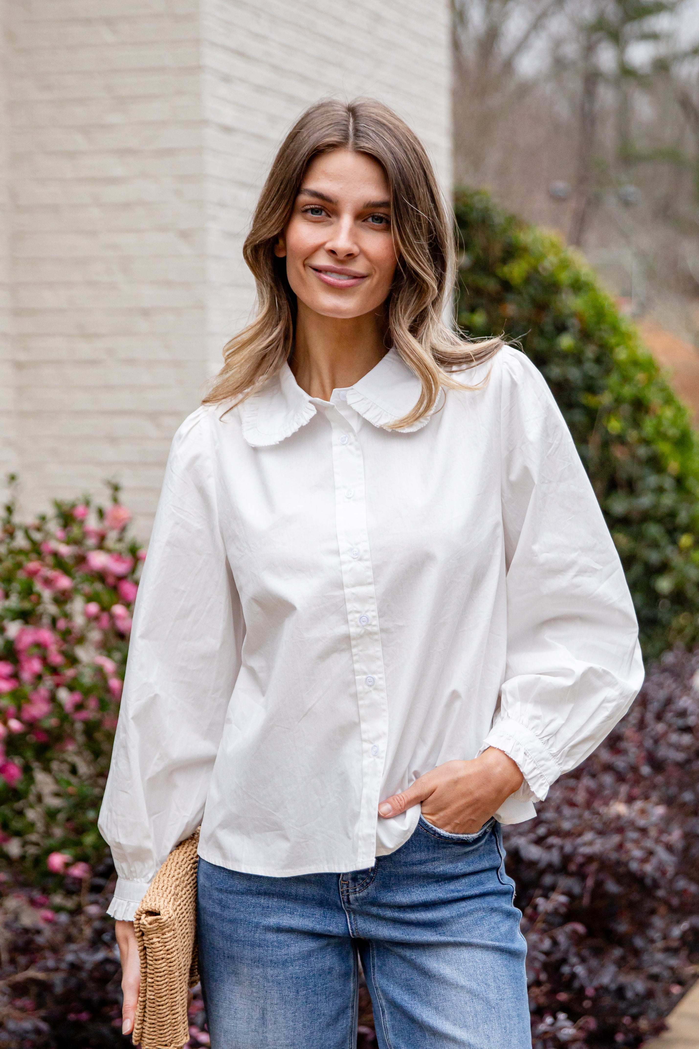 Woman wearing a white blouse and blue jeans standing outdoors with flowers and a building in the background.