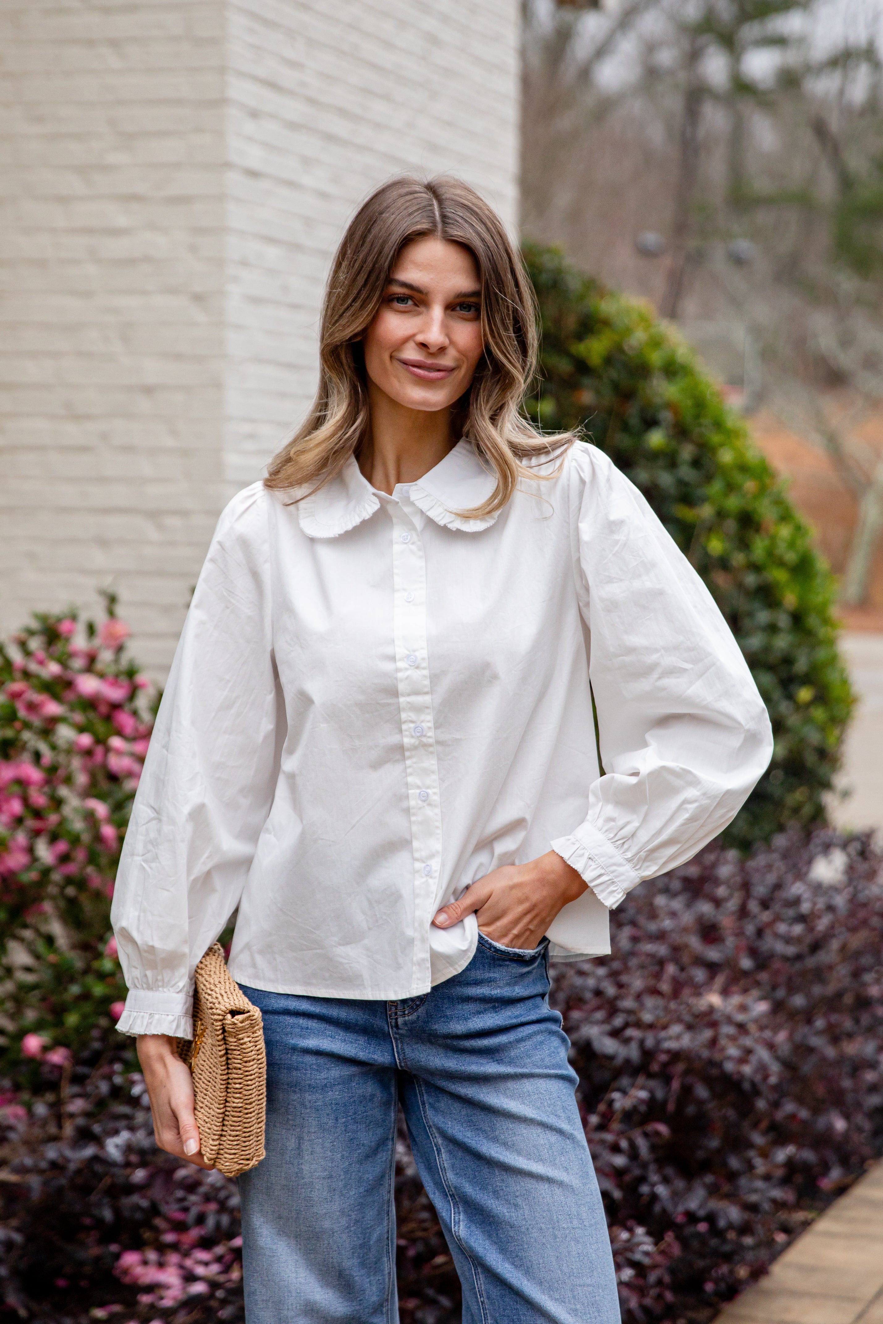 Woman wearing a white blouse and blue jeans standing outdoors with flowers and a building in the background.