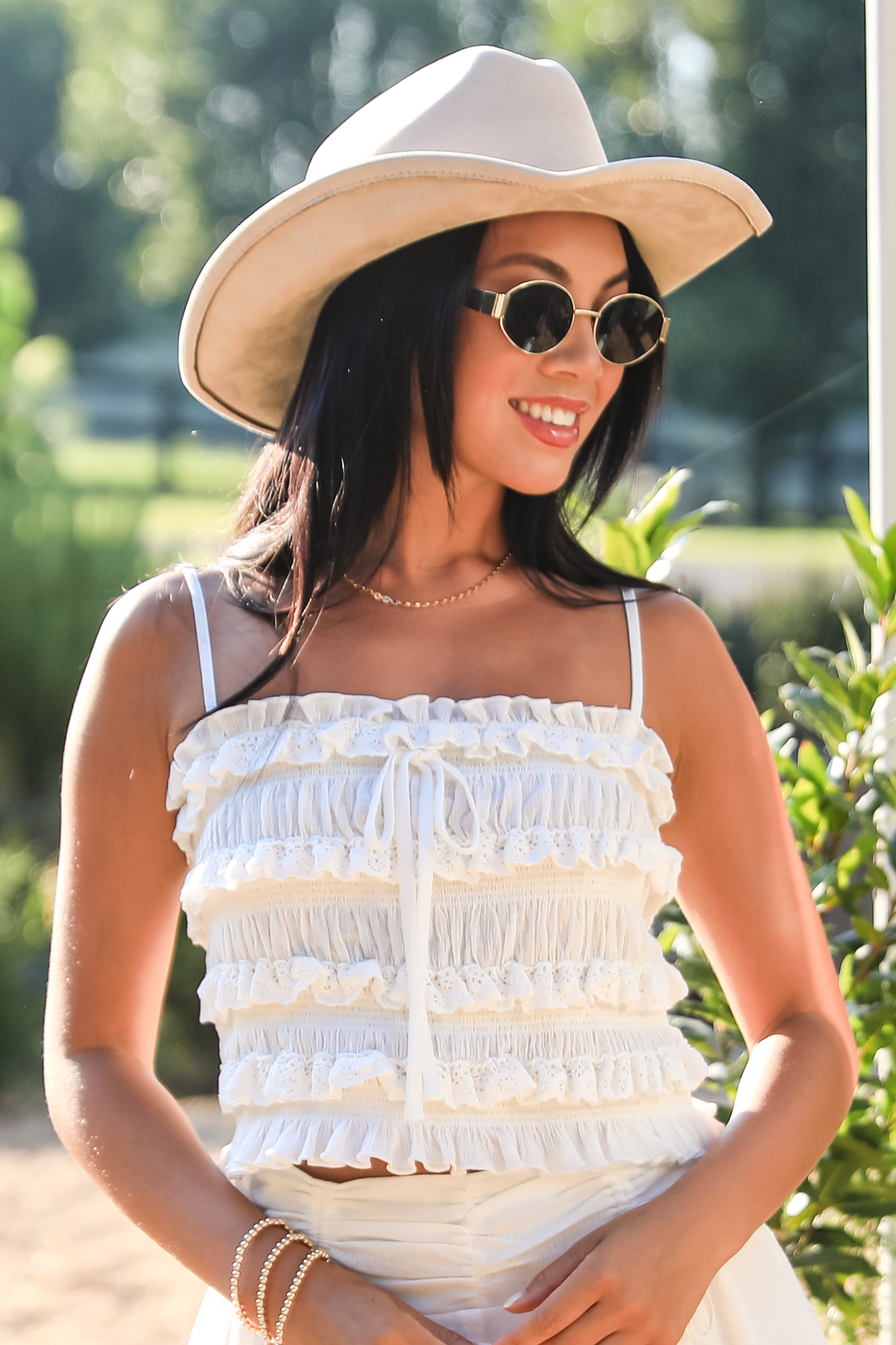 Woman wearing a white ruffled top and beige hat with sunglasses outdoors.