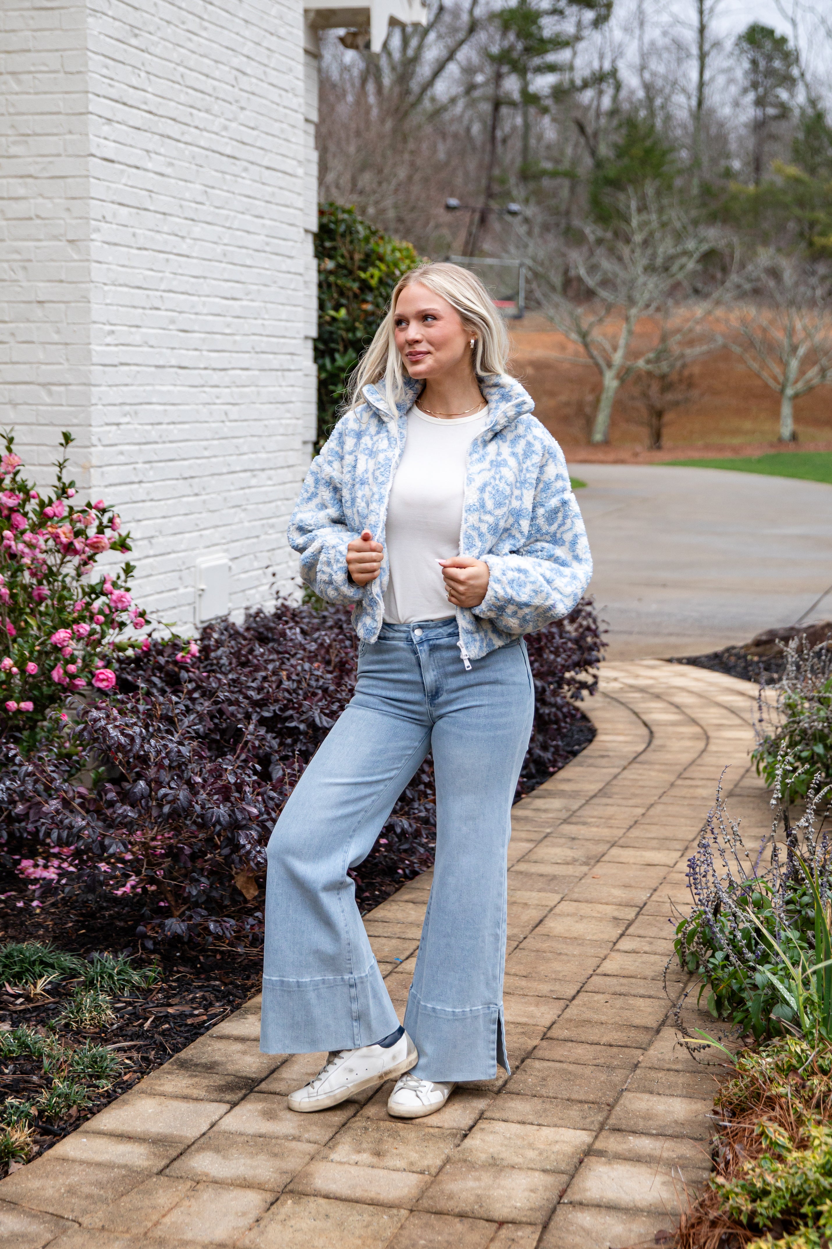Woman in a floral jacket and blue jeans standing on a brick path with a garden background