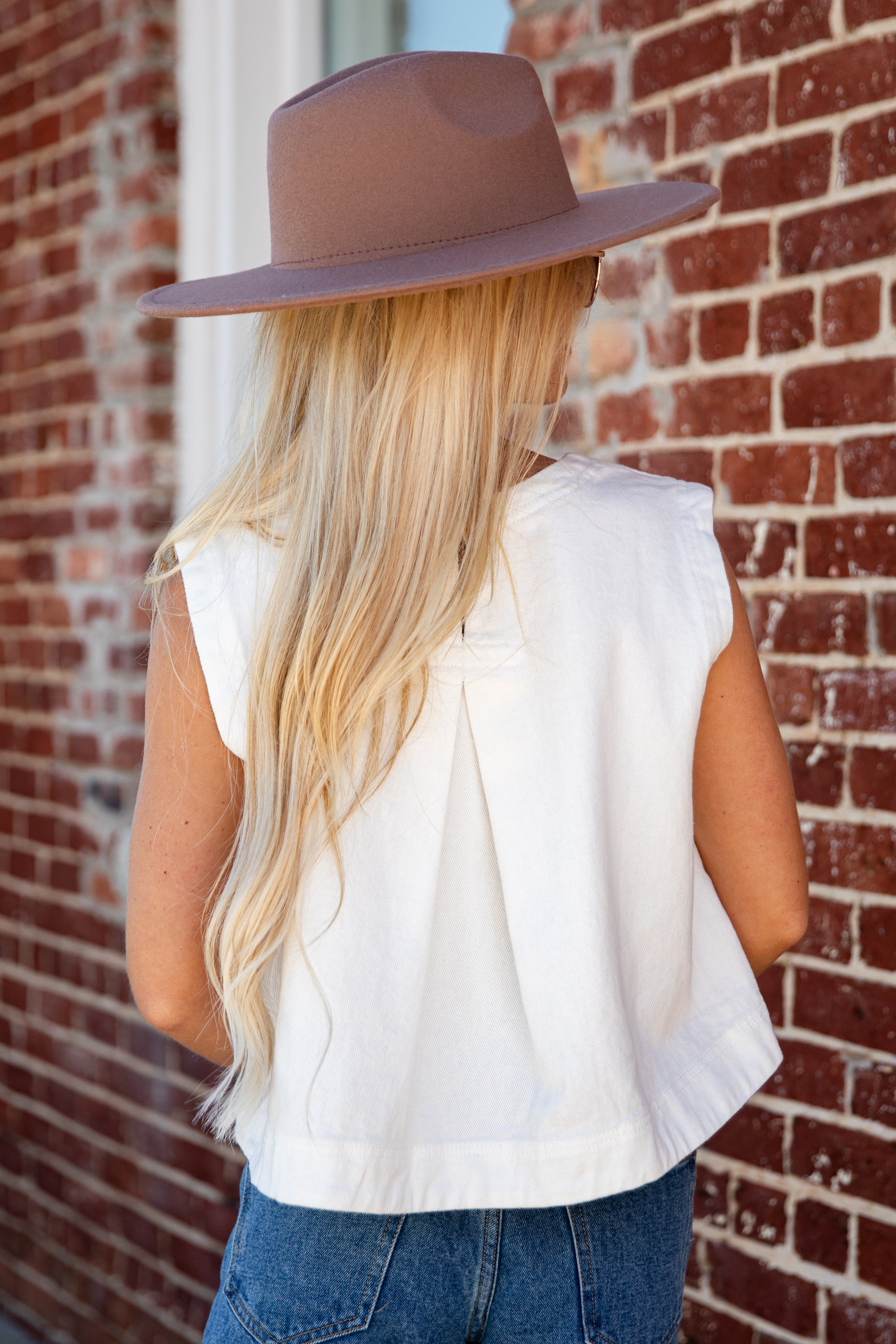 A woman with long blonde hair is shown from behind, wearing a brown wide-brim fedora hat, a white sleeveless top, and blue jeans, standing against a red brick wall.