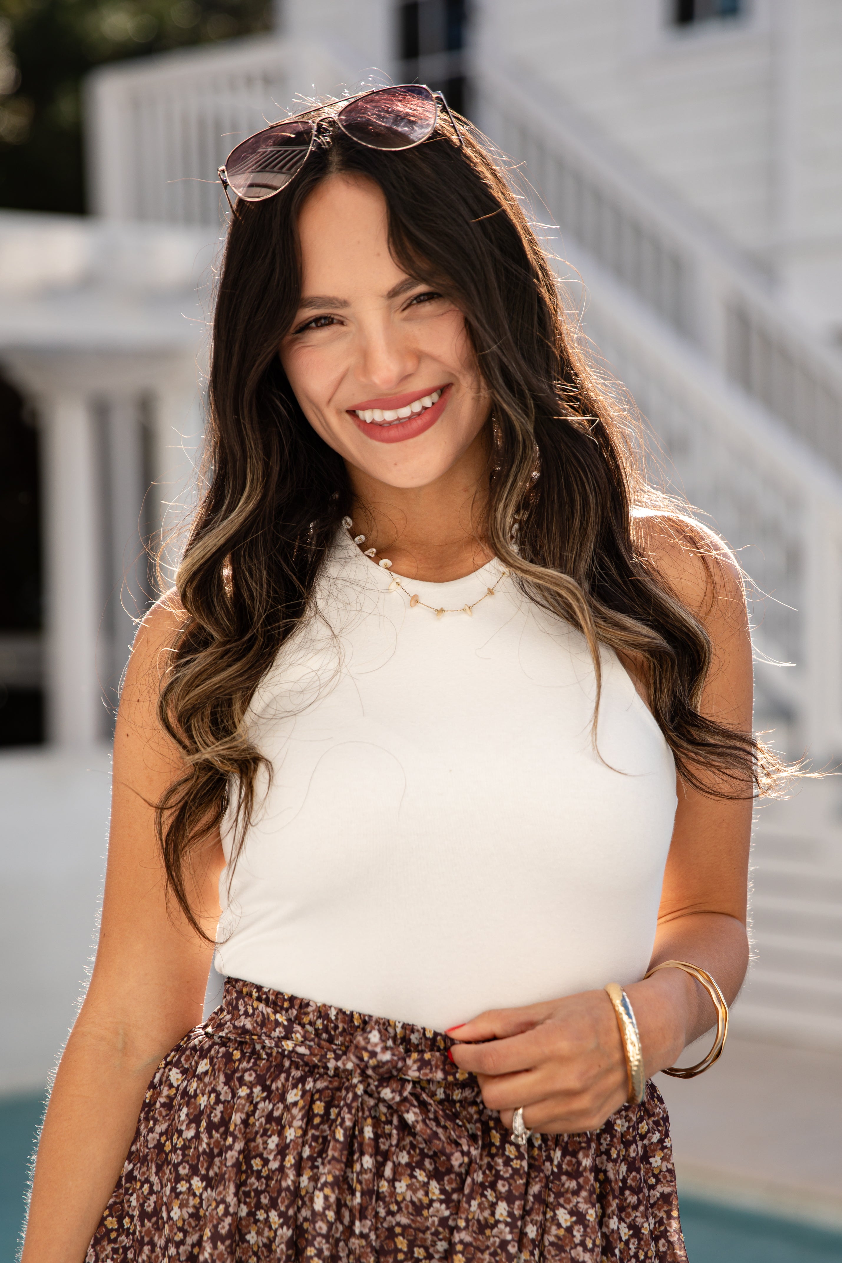 Woman wearing a white sleeveless top and patterned skirt with sunglasses on her head, standing outdoors.