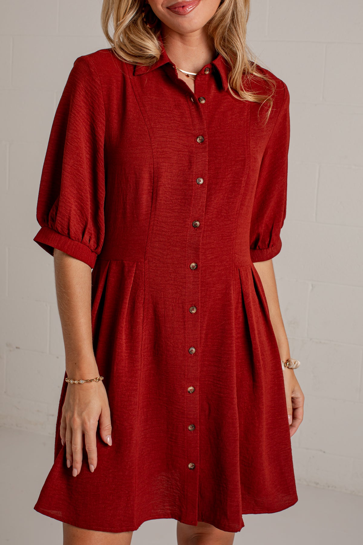 A woman wearing a red button-down shirt dress with puff sleeves and a flared skirt, standing against a light gray brick wall.