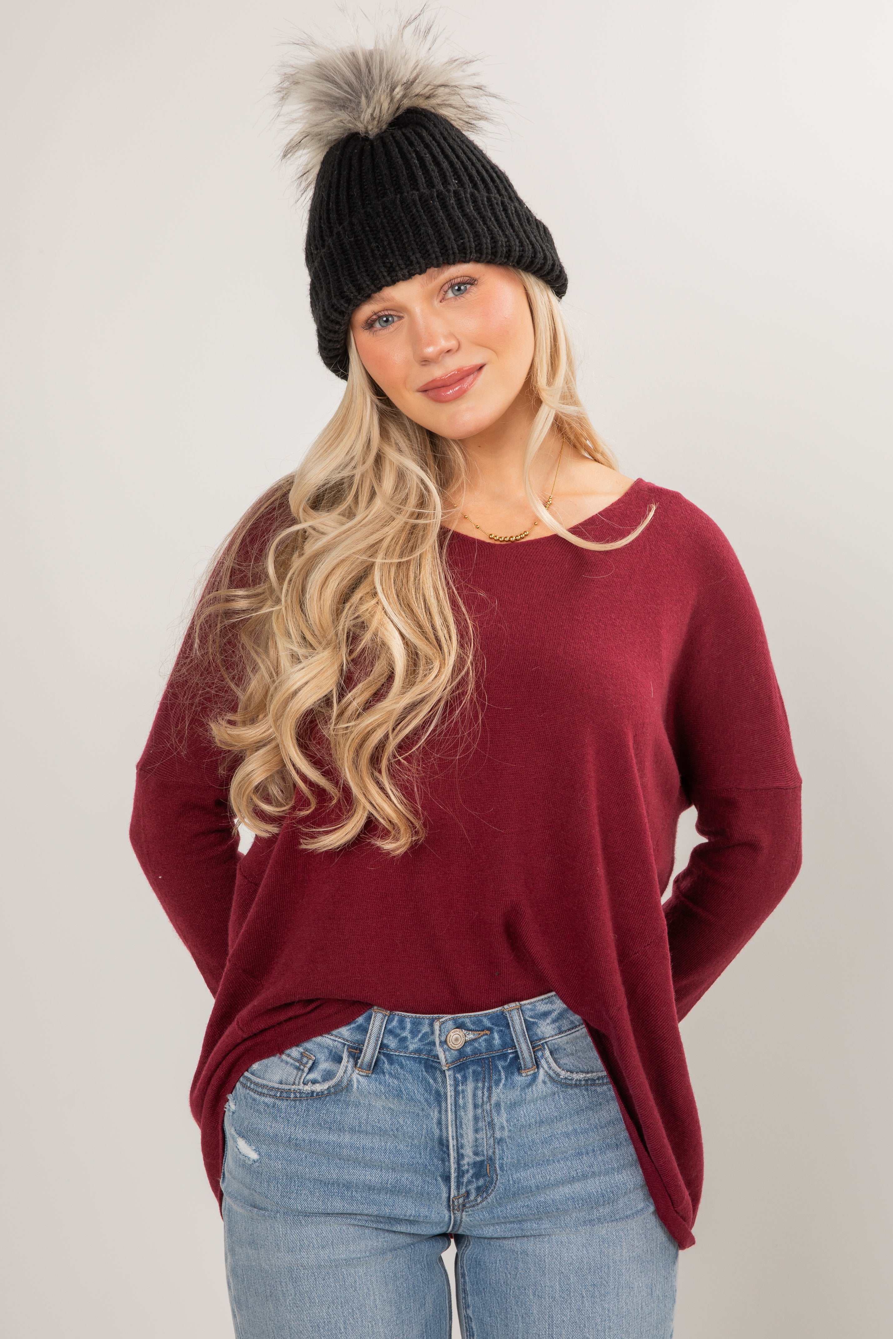 Woman wearing a burgundy sweater, black knit beanie with pom-pom, and blue jeans against a plain background