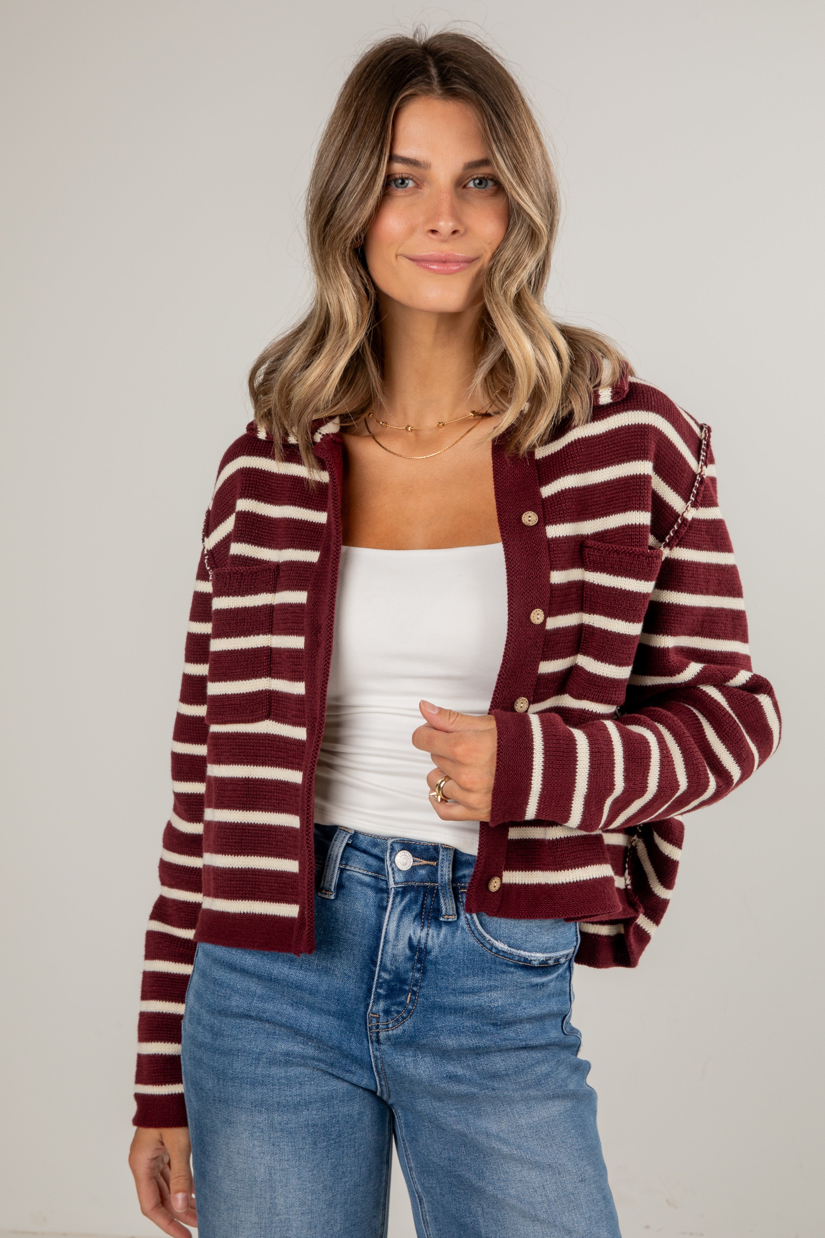 Woman wearing a maroon and white striped cardigan over a white top and blue jeans on a plain background