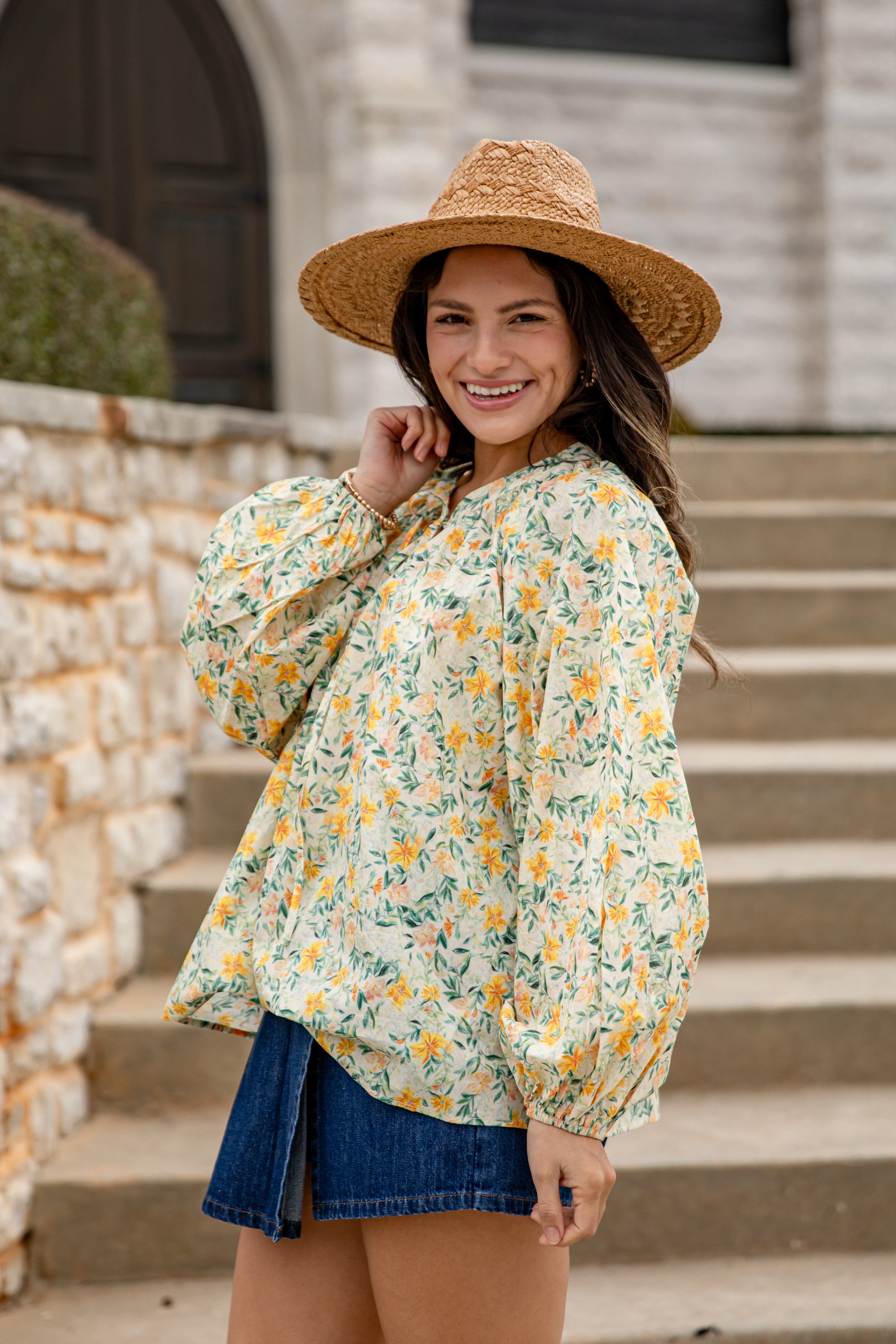 Woman wearing a floral blouse and straw hat standing on steps outdoors.