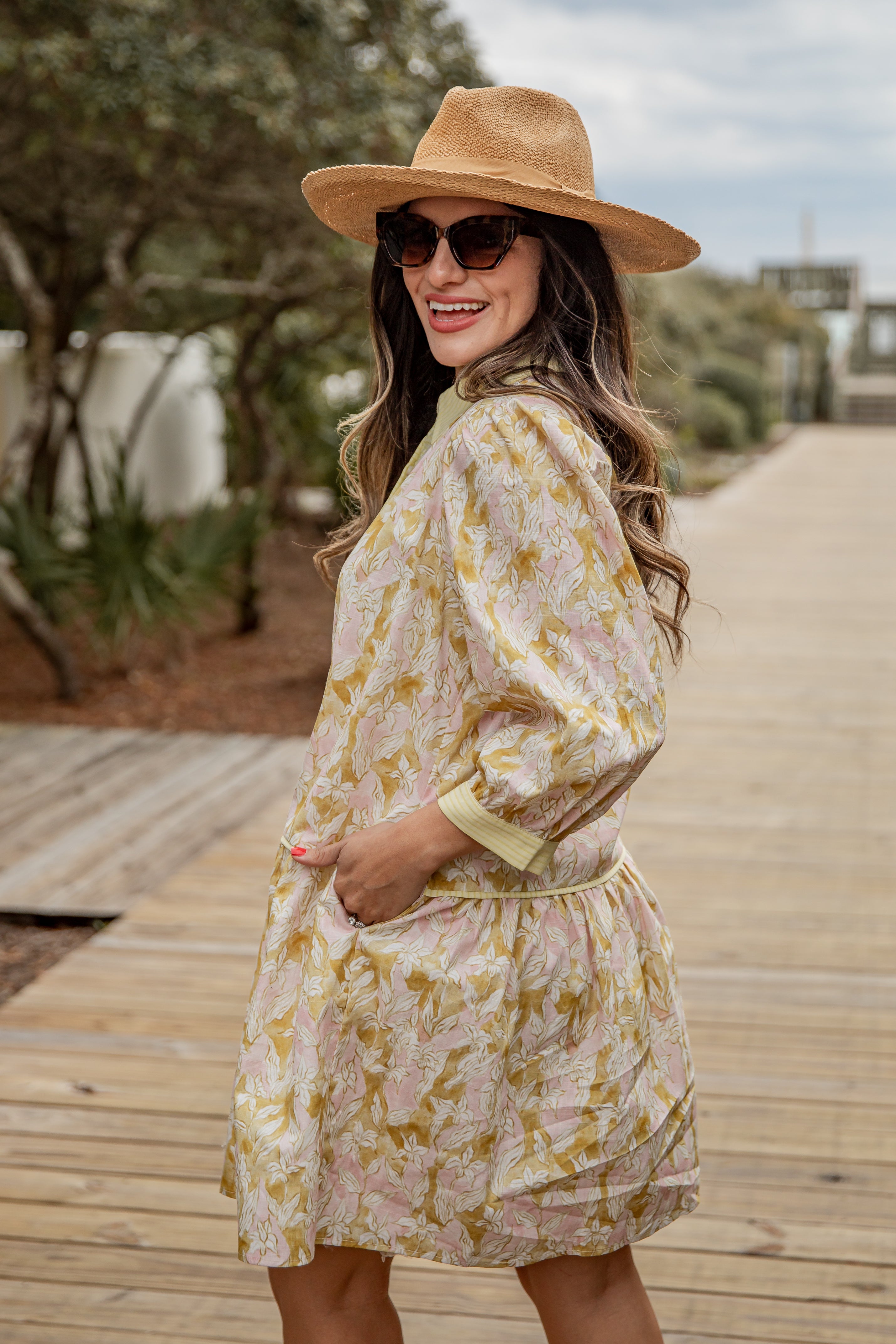 Woman in a floral dress and hat standing on a wooden path with greenery in the background