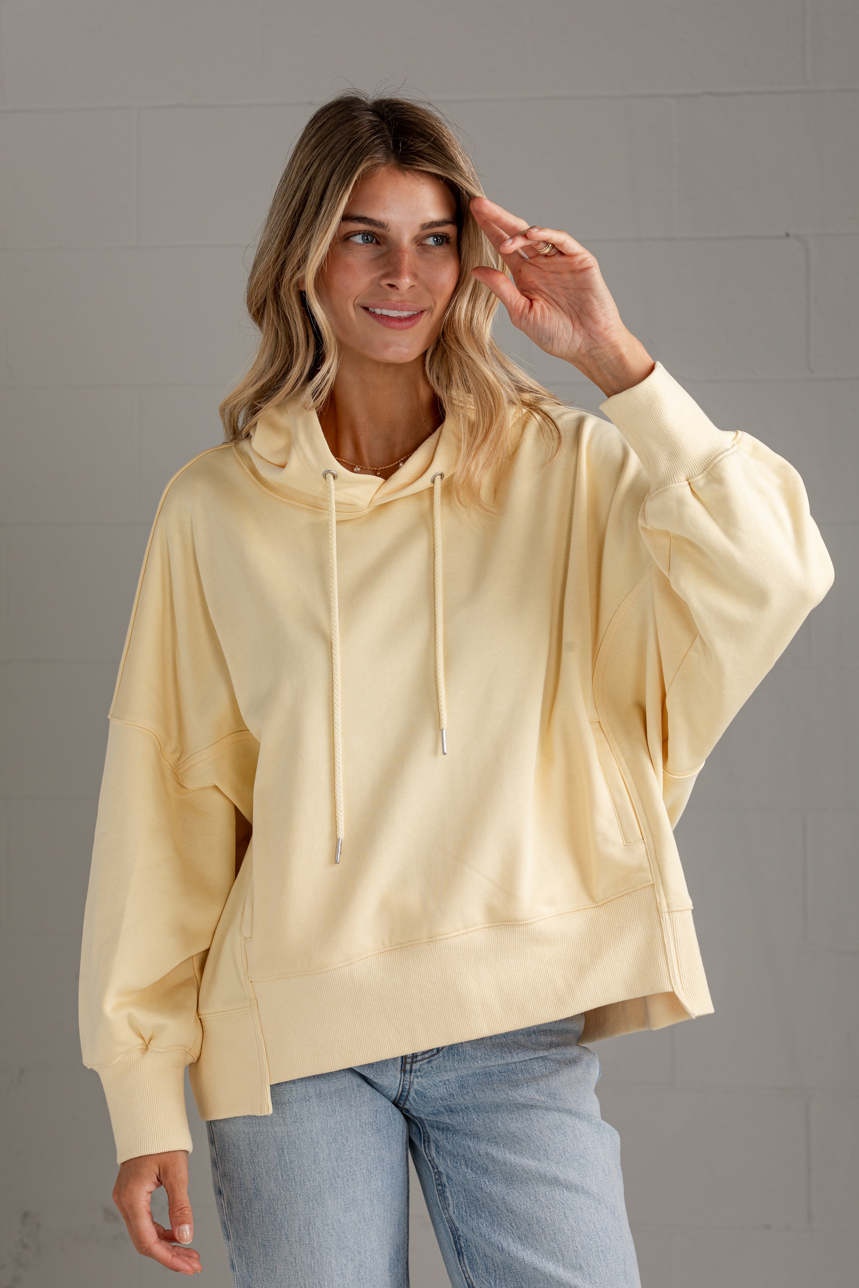 A woman wearing a pale cream-colored oversized pullover hoodie with balloon sleeves and drawstrings, paired with light blue jeans, standing against a gray brick wall.
