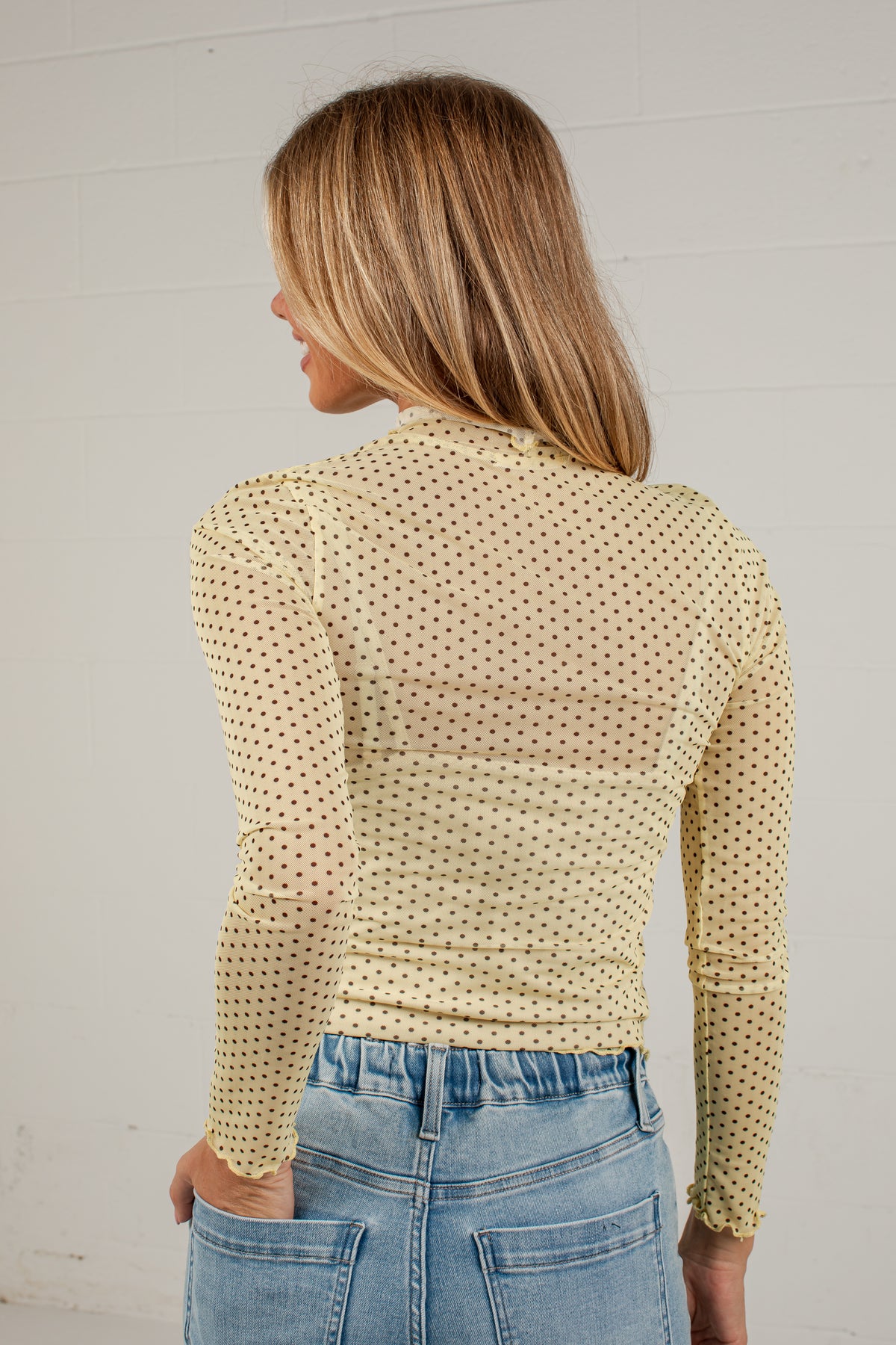A woman shown from the back wearing a pale yellow long-sleeve top with a small repeating dark polka dot pattern, paired with light blue jeans, against a white brick wall.