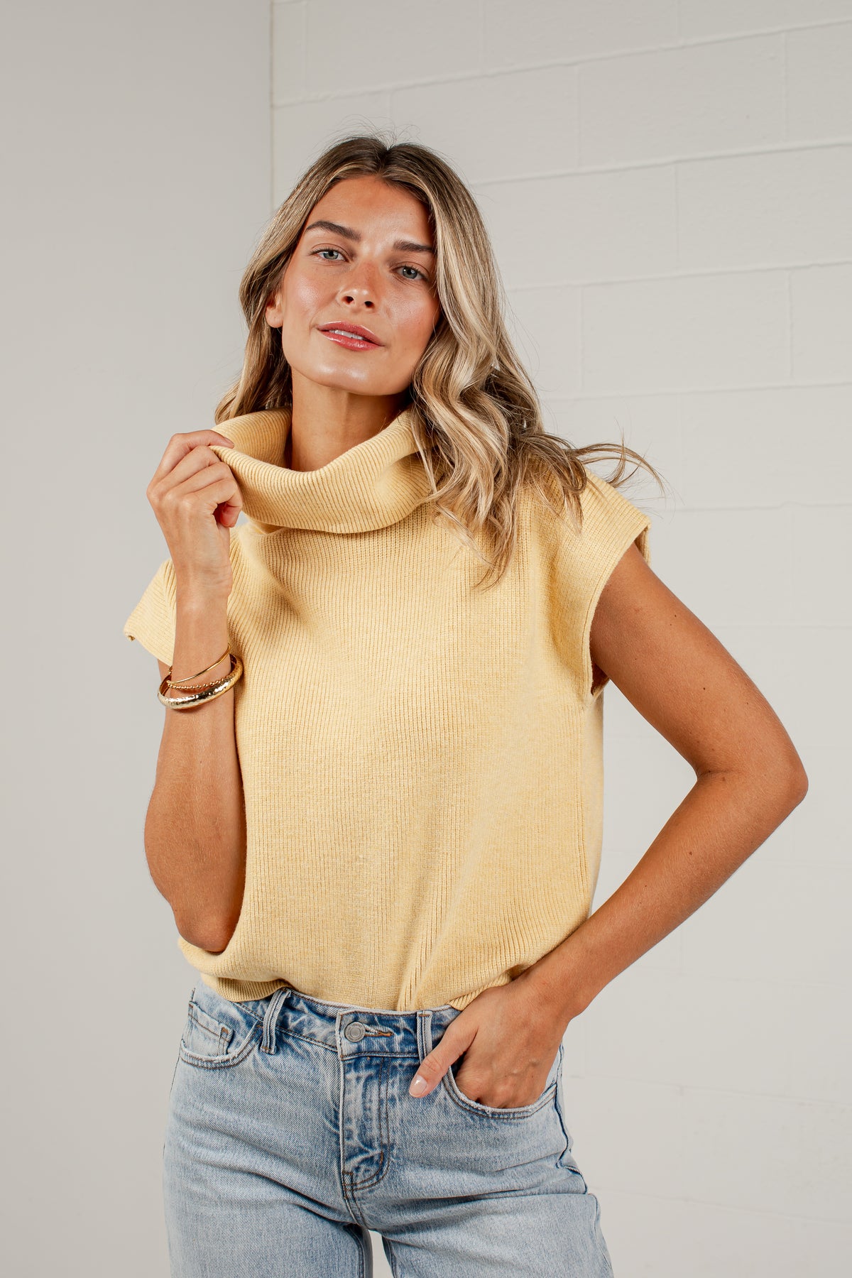 A woman wearing a pale yellow ribbed knit mock neck top with cap sleeves and light-wash blue jeans, standing against a white brick wall.