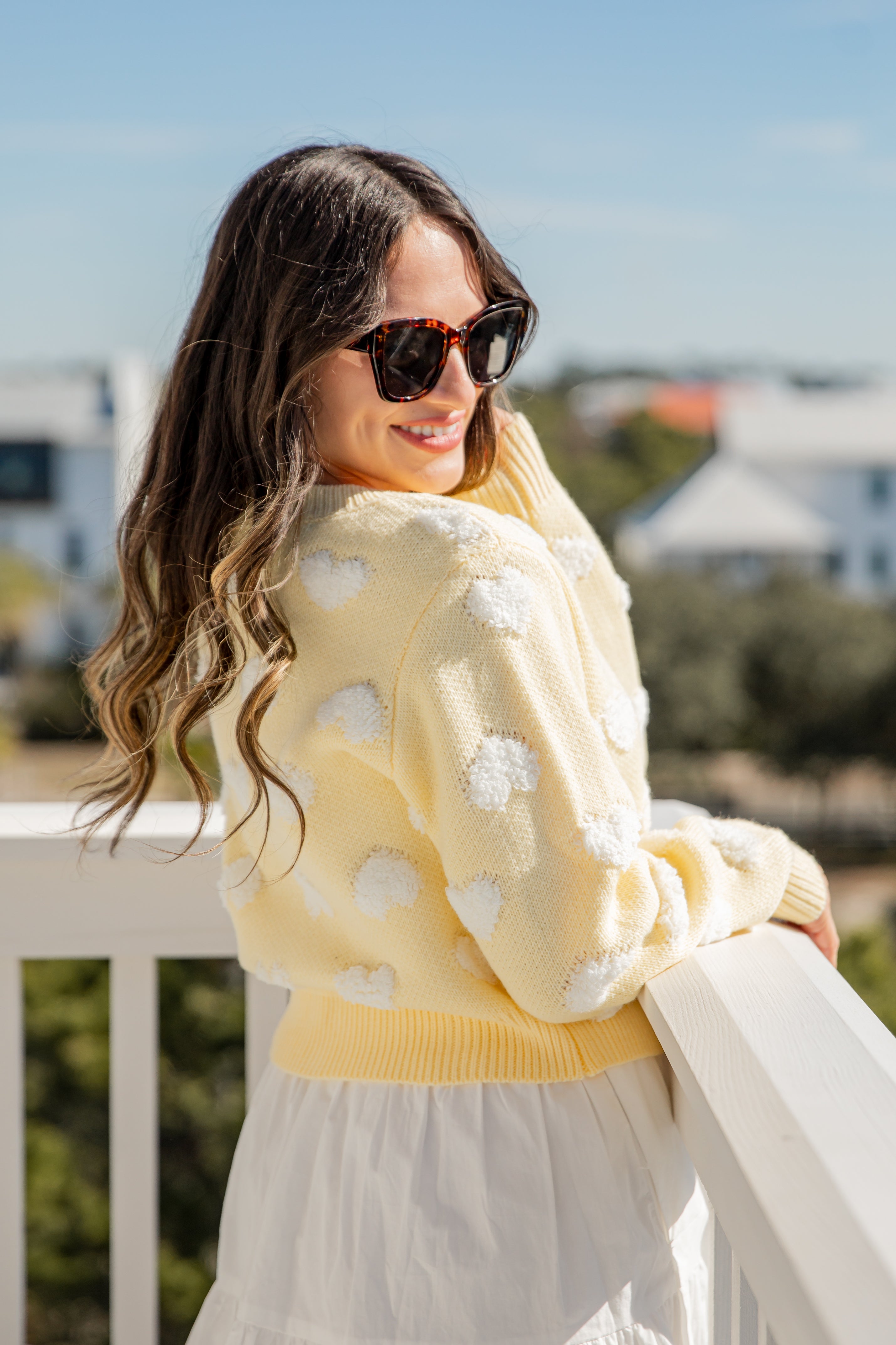 Woman wearing a yellow cardigan with white floral patterns, sunglasses, and holding a white scarf outdoors.