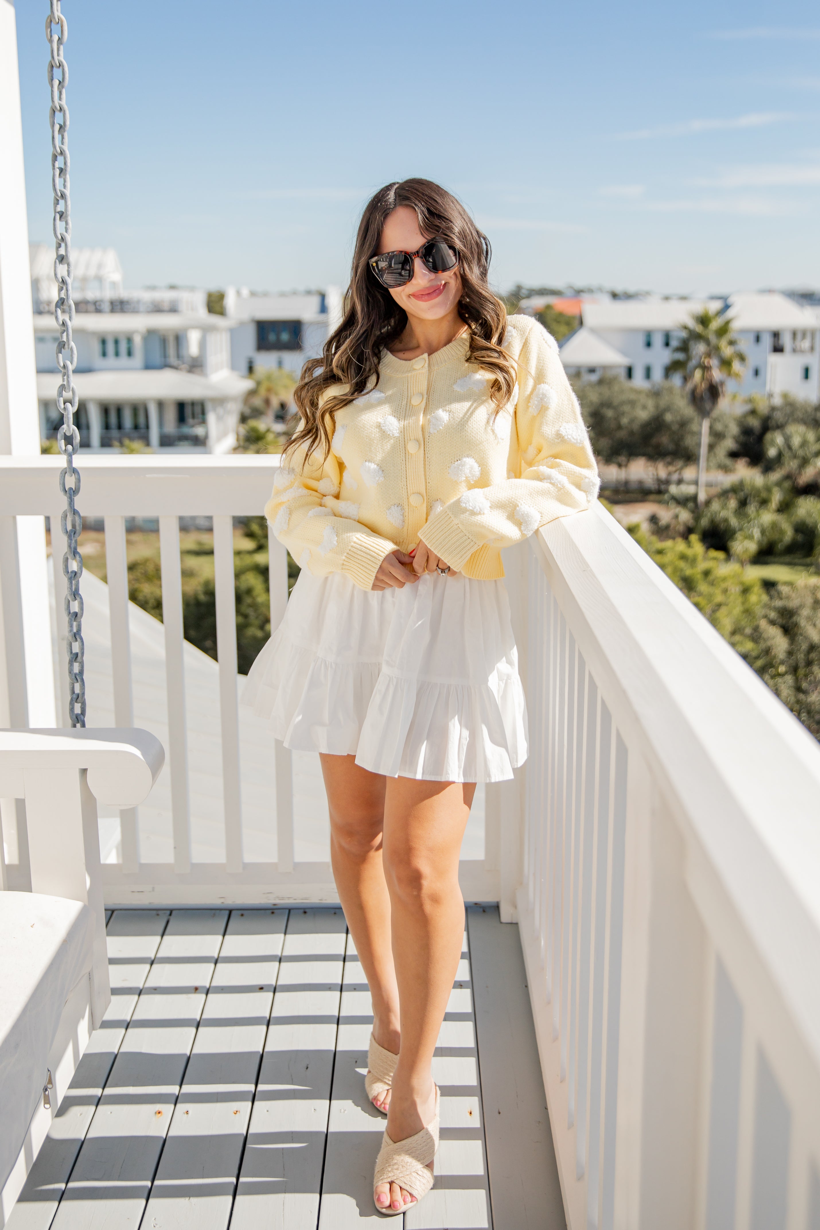 Woman in a yellow jacket and white skirt standing on a balcony with a scenic background.