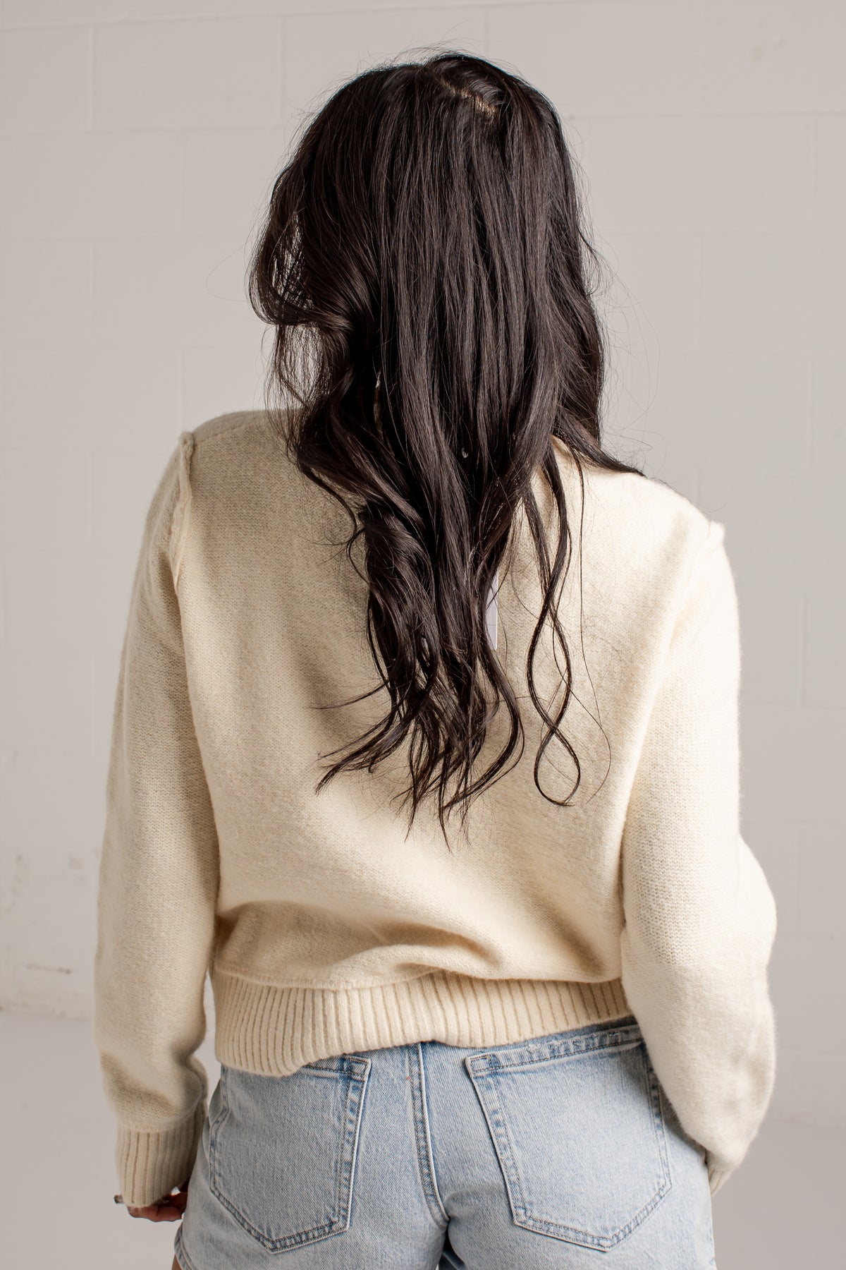 A woman viewed from behind wearing a cream-colored knit sweater and light blue jeans, with her long dark hair visible.