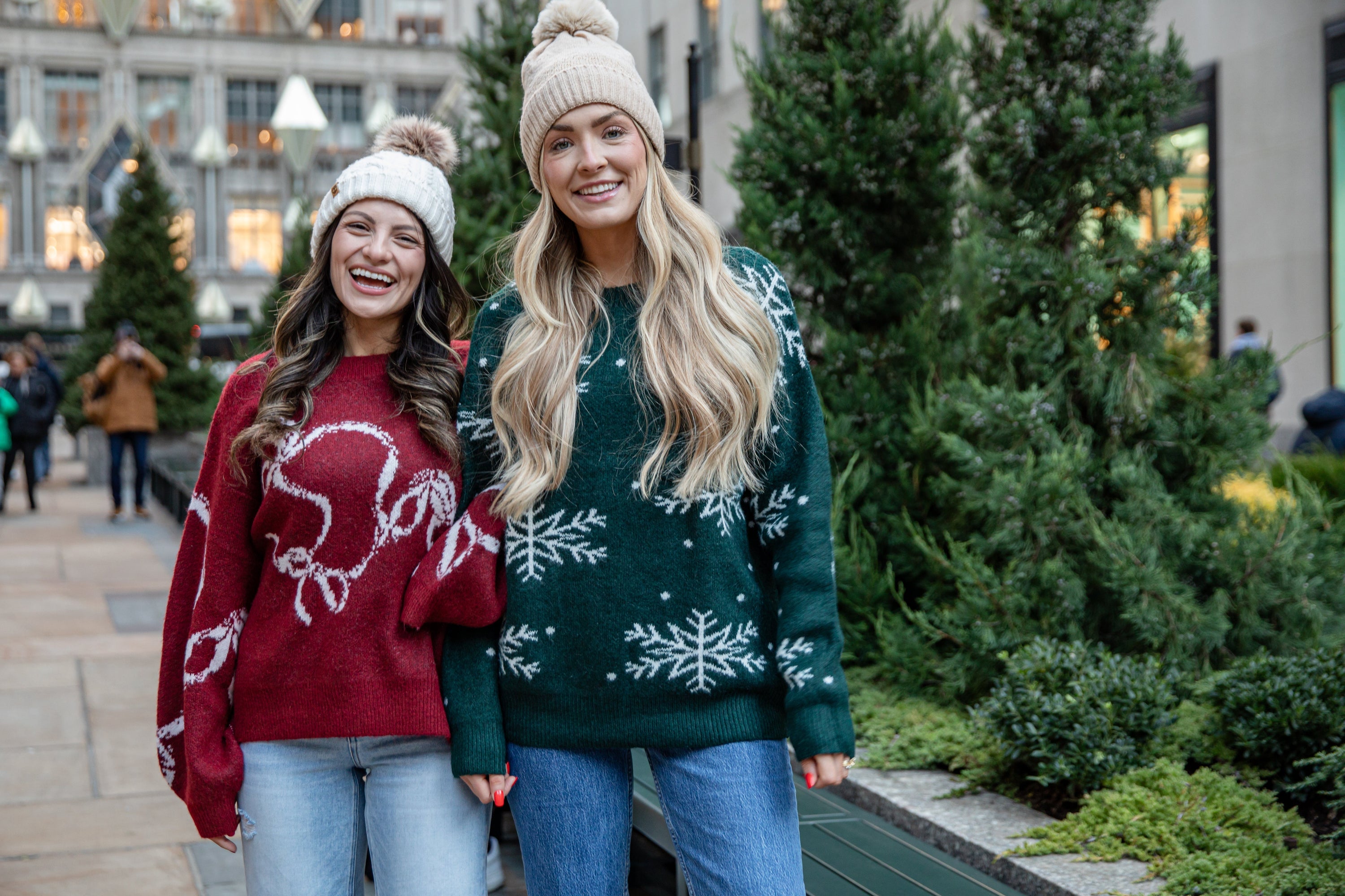 Two women smiling outdoors near evergreen trees, wearing winter knit sweaters and beanies. One wears a red sweater with a white holiday graphic and light-wash jeans, while the other wears a dark green sweater with white snowflake details and blue jeans.