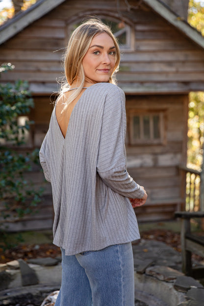 Woman wearing a light gray open-back cardigan and blue jeans standing in front of a wooden cabin.
