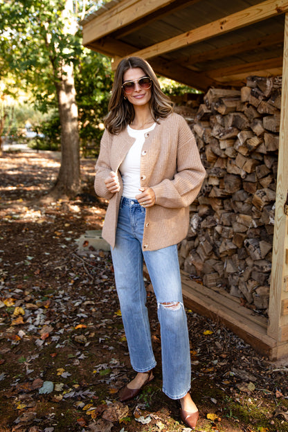 Woman standing outdoors near a stack of firewood, wearing a beige cardigan, white shirt, blue jeans, and sunglasses.