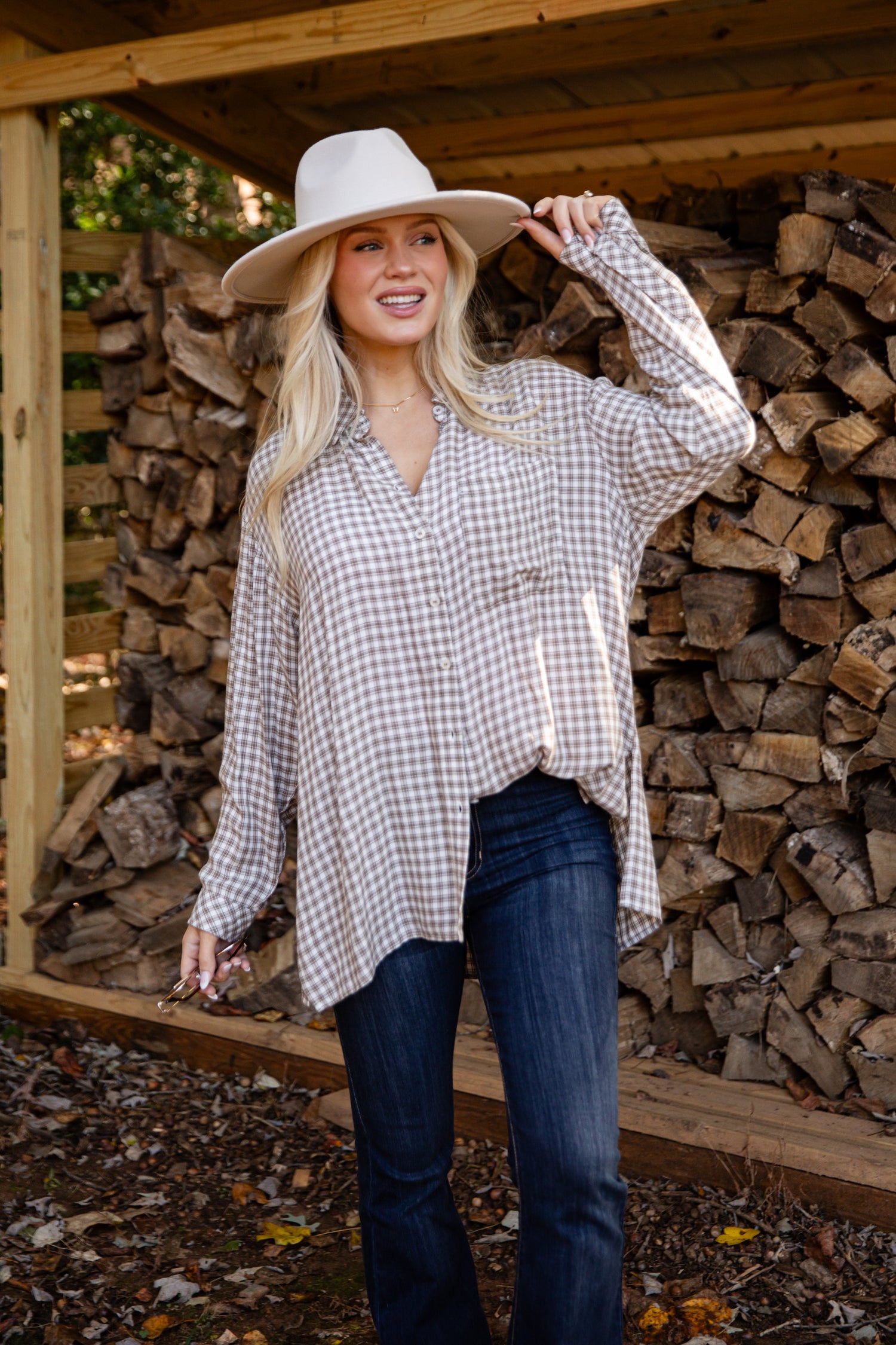 Woman wearing a checkered shirt and cowboy hat standing in front of stacked firewood.