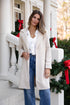 Woman in a beige coat standing on a decorated porch with Christmas decorations.